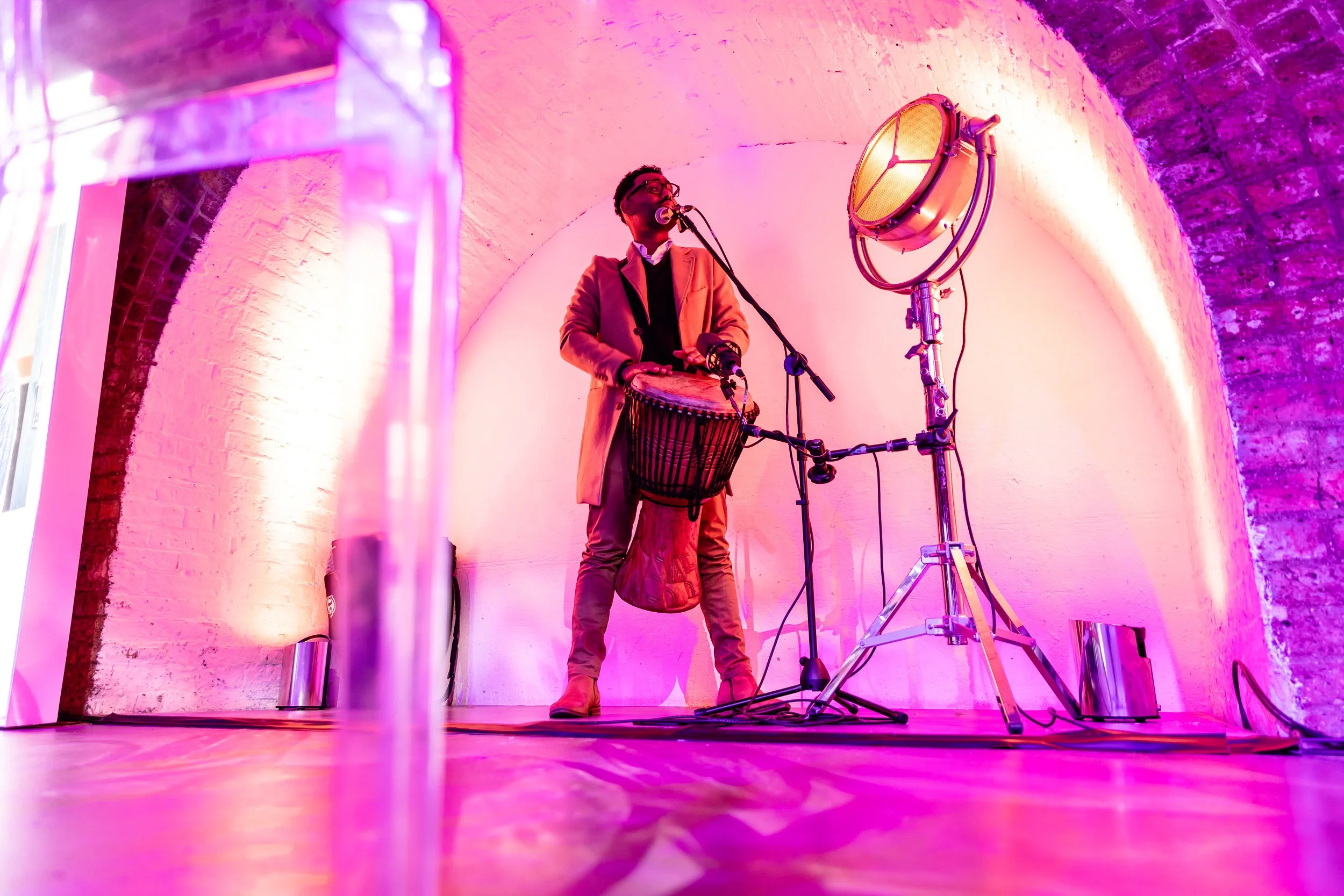 Musician playing a djembe drum under colorful stage lights in an arched, brick-walled venue.