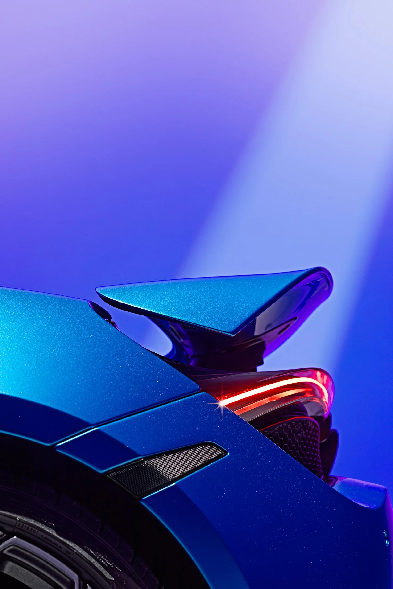 Close-up of a blue sports car's rear spoiler and taillight on a purple gradient background.