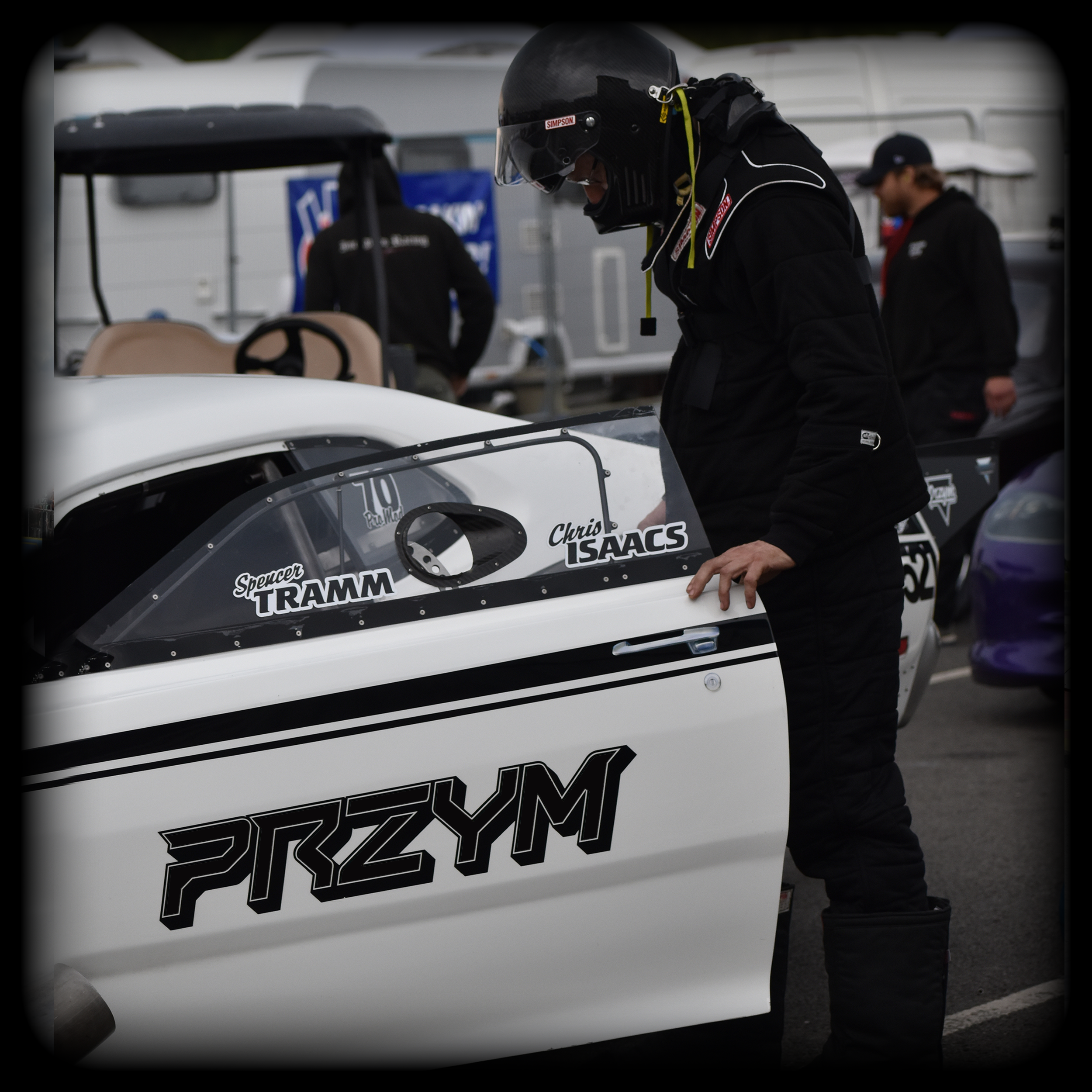 A race car driver in black racing gear and helmet standing next to a white open-cockpit race car with PRZYM sponsor decals. The background features trailers and another person in black clothing.