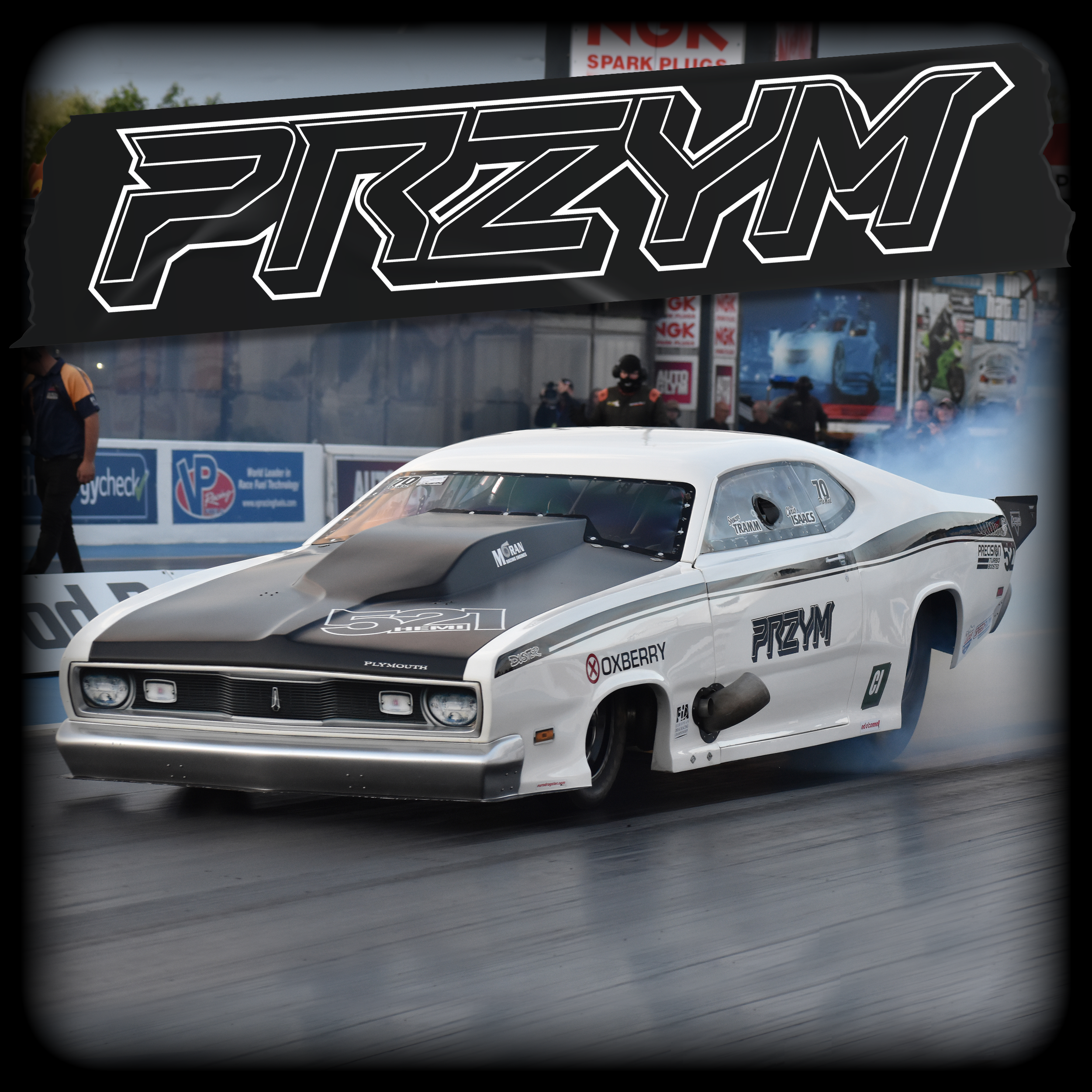 A white Plymouth Duster Pro mod with black accents speeding down the track, with a large illuminated sign reading 'PRZYM' in black and white above it, and group of people, some wearing racing uniforms, watching in the background.