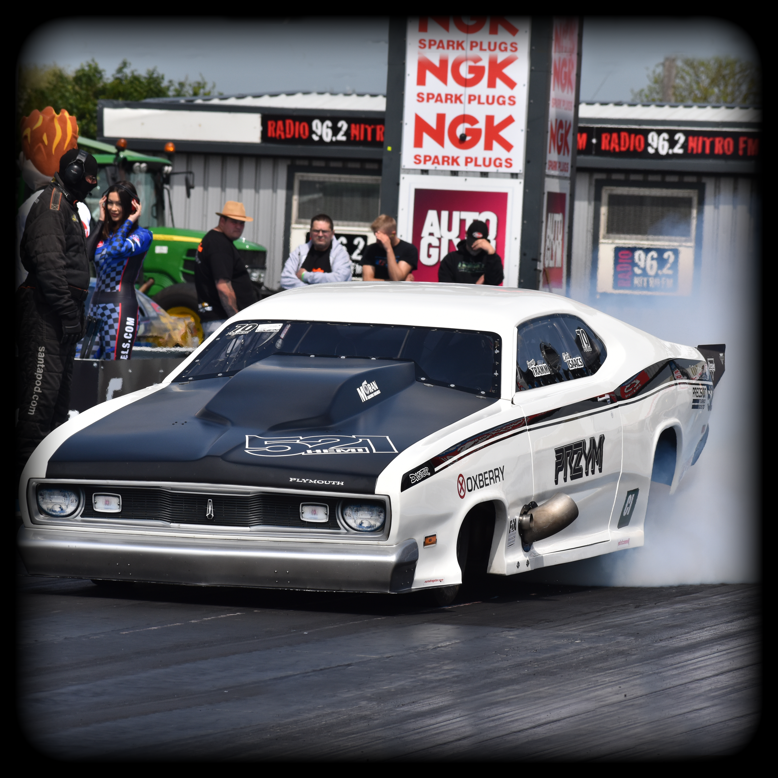 A high-performance Plymouth Duster Pro mod race car on a drag strip with people standing nearby, some wearing racing gear and headphones, and signage advertising NGK spark plugs and radio station 96.2 FM.