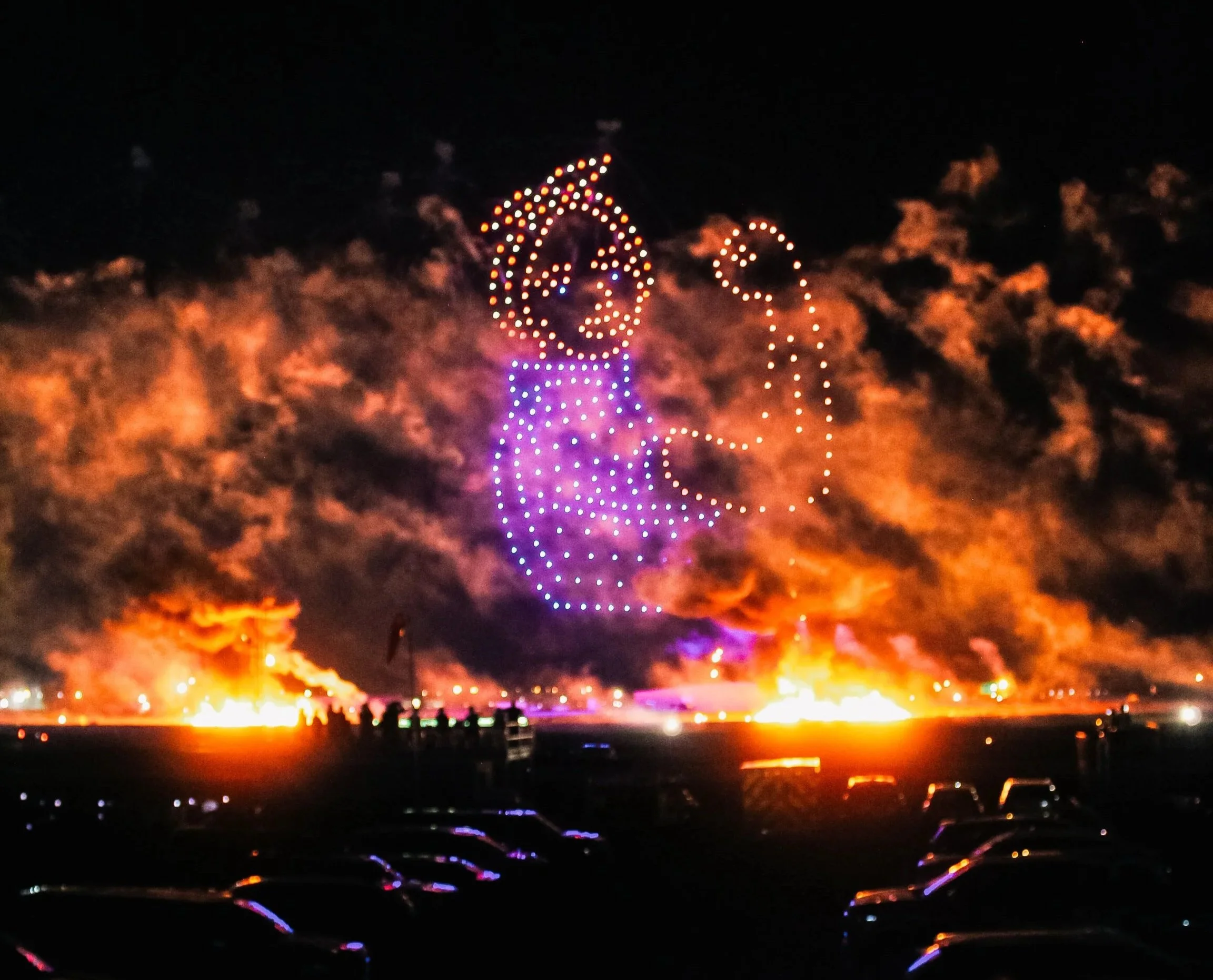 A night scene of a fireworks display featuring an drone light show against a background of exploding fires and thick smoke.