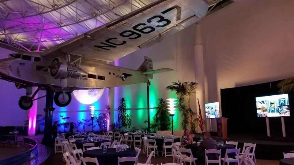 Indoor event space featuring a vintage aircraft hanging from the ceiling, with colorful lighting and tables set up for dining, including a small stage with two screens.