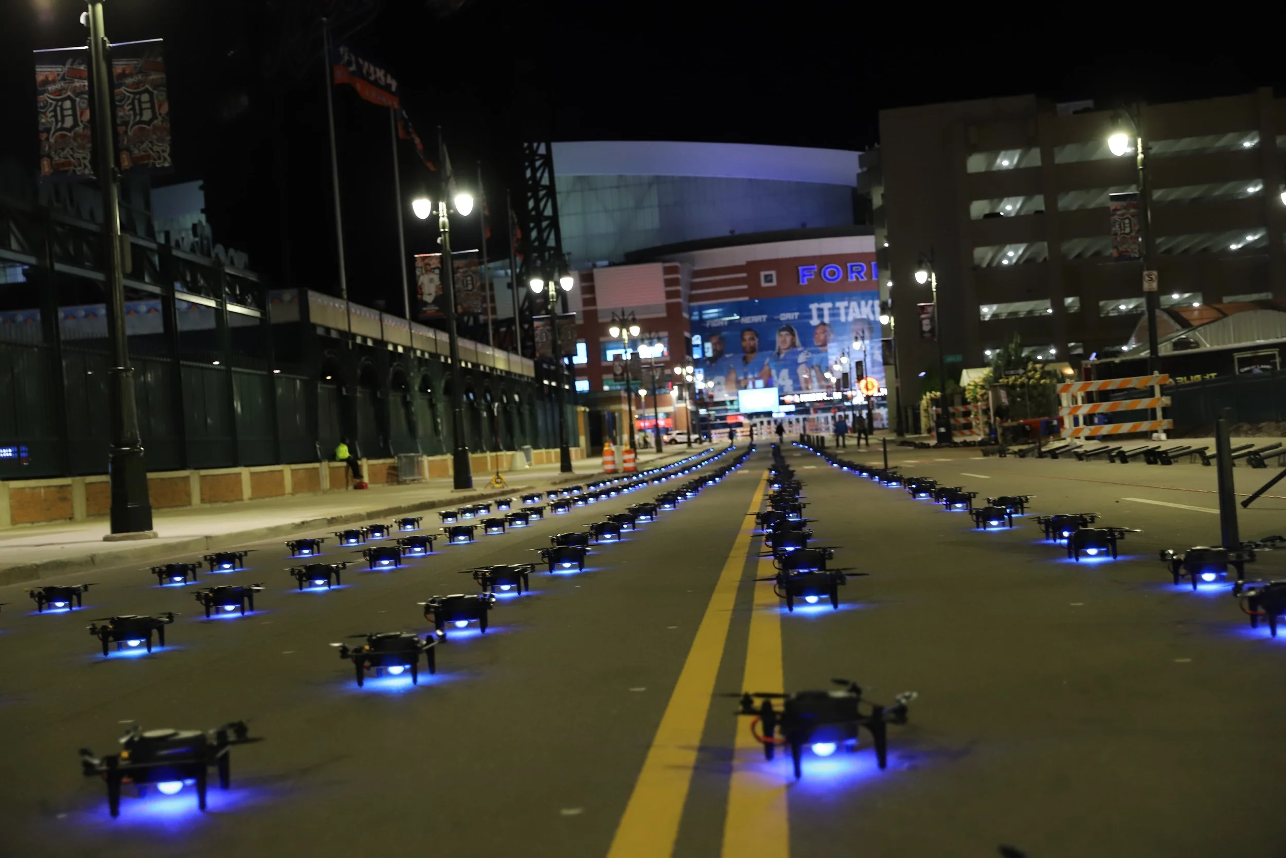 Row of small drones with blue lights on a city street at night, with buildings and signs visible in the background.