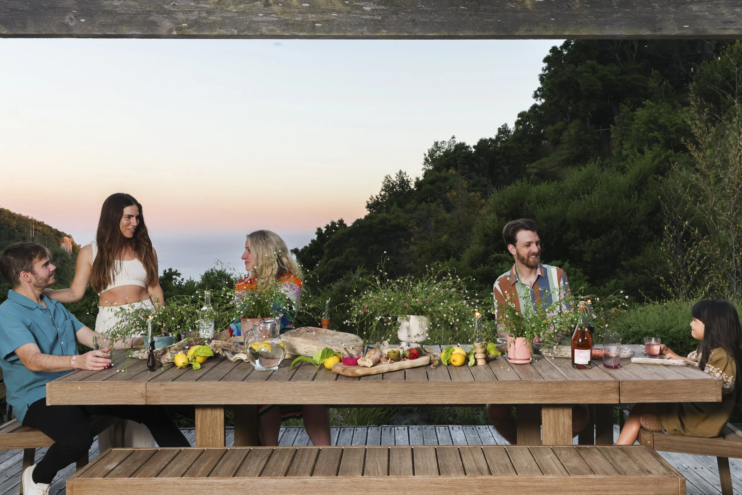 Outdoor dining scene at sunset with friends gathered around a wood table overlooking the Big Sur coastline, styled by CCG Designs.