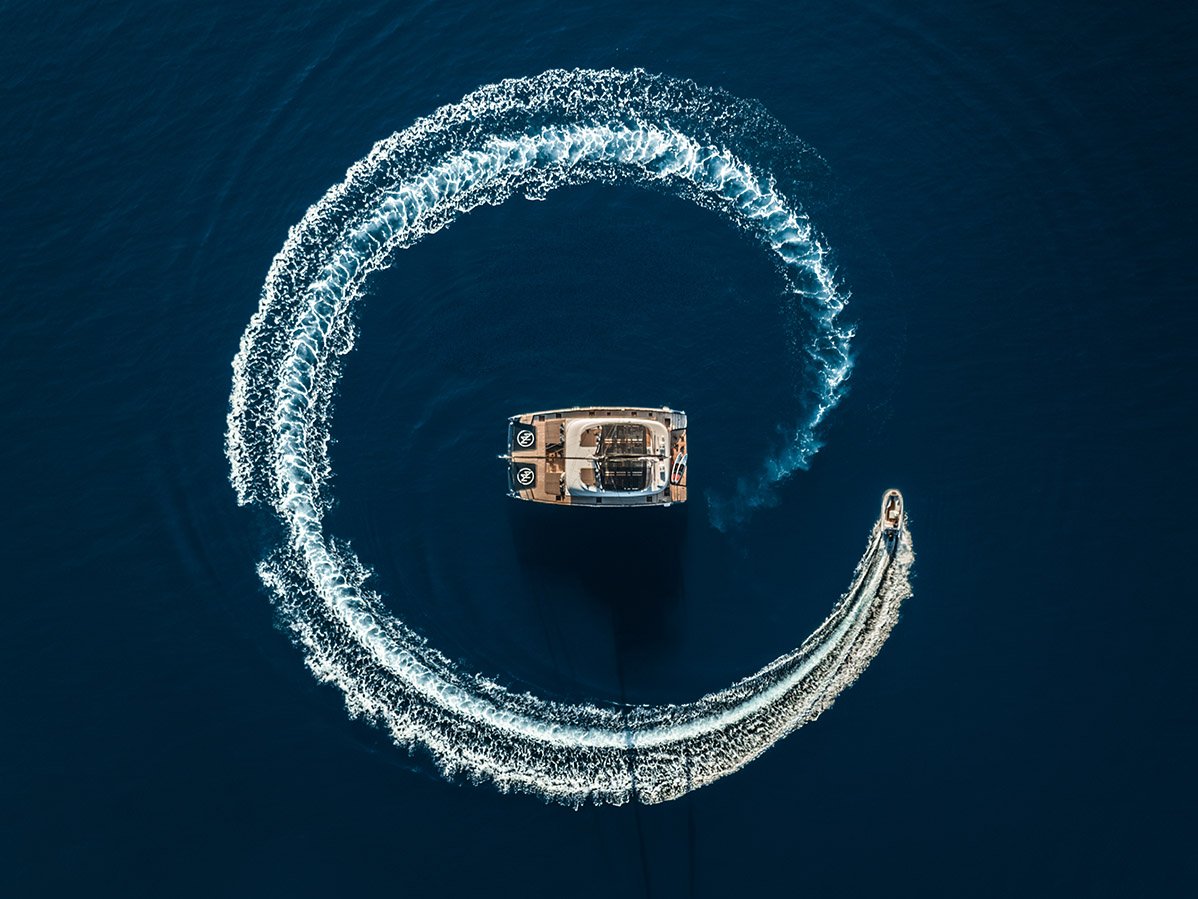 Luxury charter lifestyle cinematography from Greece: An aerial drone shot capturing a high-speed tender creating a circular wake around a stationary power catamaran in the deep blue Ionian Sea.
