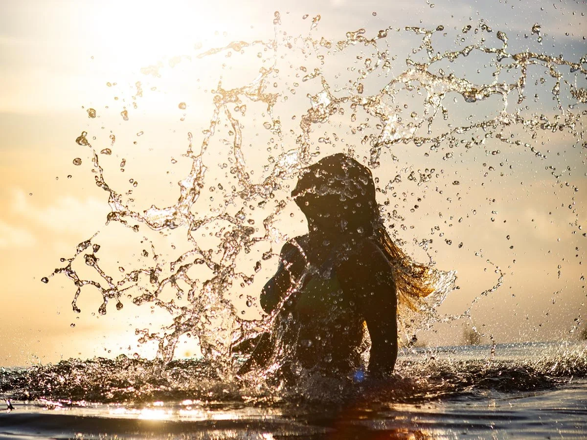 Luxury hospitality cinematography from Greece, capturing a candid, back-lit moment of a guest emerging from the water at sunset with dynamic water spray, showcasing the aspirational Mediterranean lifestyle.