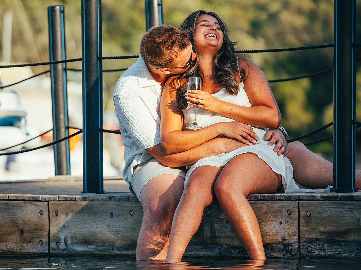 Candid destination engagement photography in Greece: A couple celebrating on a wooden jetty at sunset with champagne, captured by Ionian lifestyle photographer Joe Snowdon.