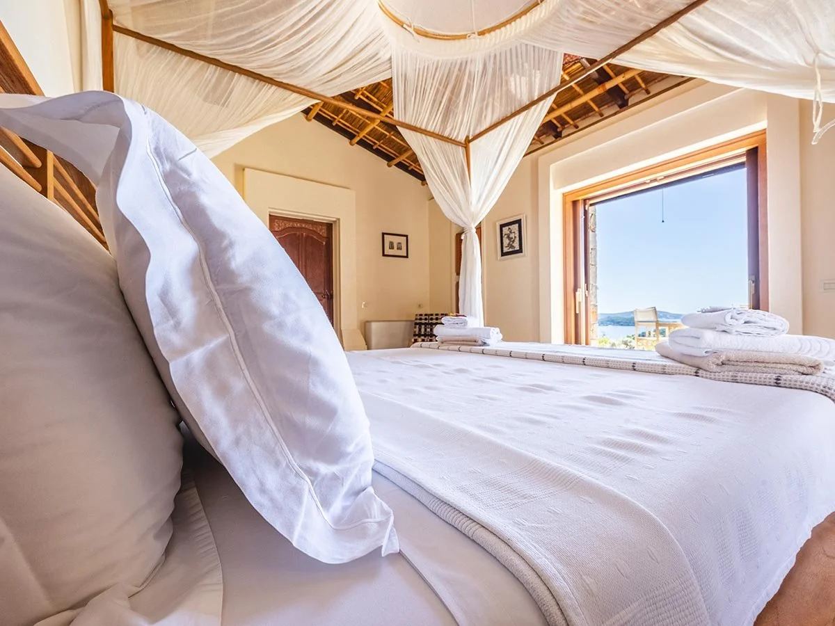 Professional wide-angle interior photography of a luxury villa bedroom in Greece, featuring a four-poster bed with canopy, exposed bamboo ceiling, and an open balcony overlooking the Ionian Sea.