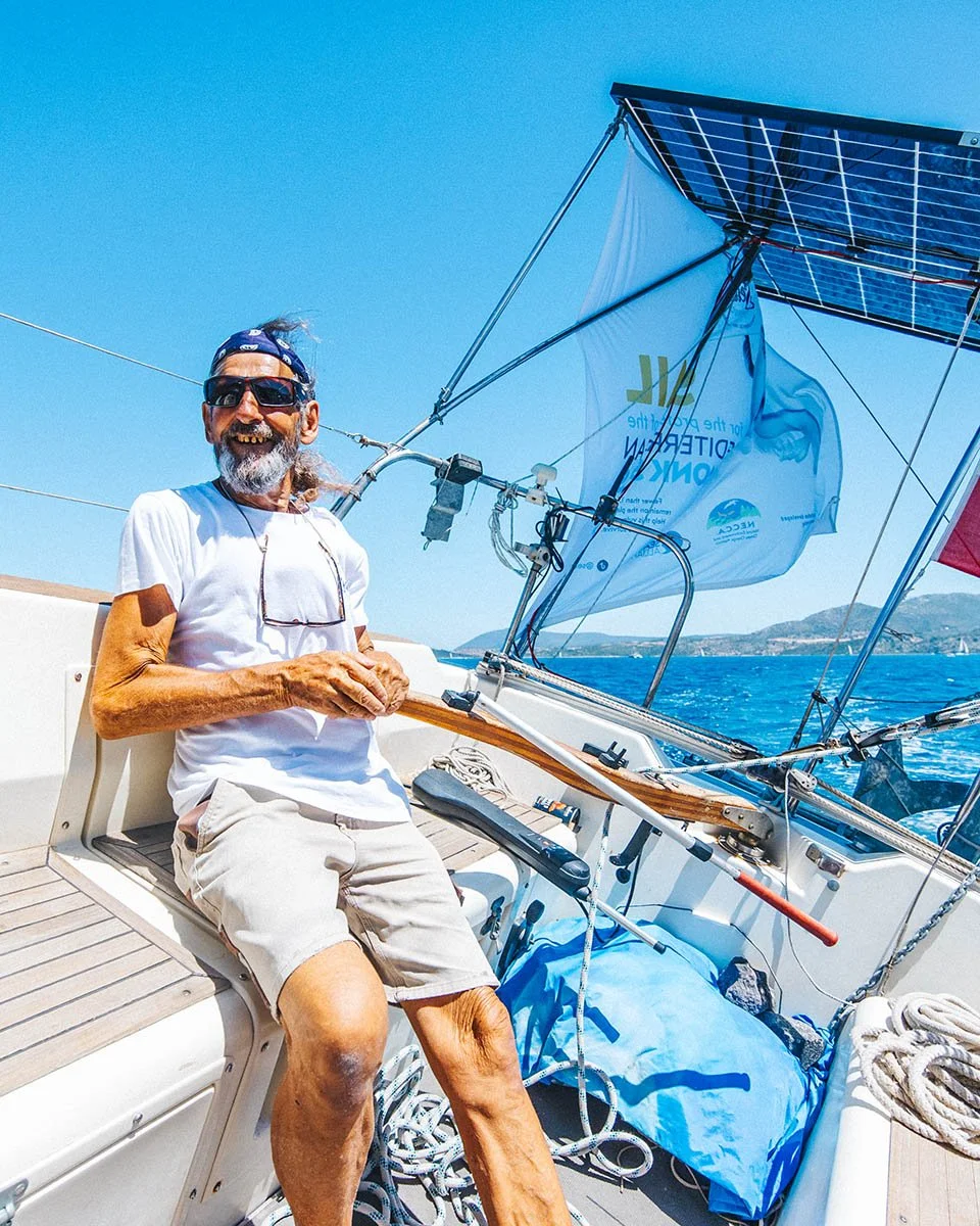 Authentic sailing lifestyle portrait of a Greek skipper at the helm of a yacht in the Ionian Sea, featuring solar panels and marine conservation branding.