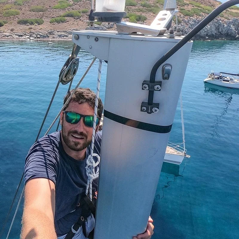 Professional yacht videographer and qualified skipper Joe Snowdon at the top of a sailboat mast in Greece, demonstrating the technical climbing and rigging skills required for high-angle maritime cinematography.