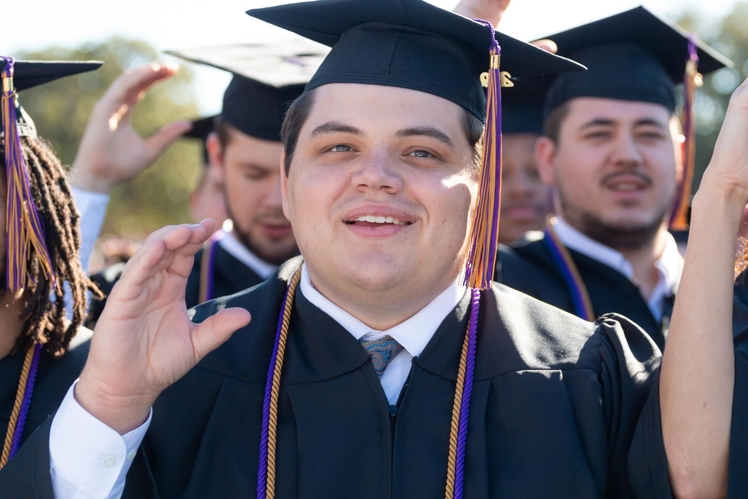 Group of diverse graduates in caps and gowns during graduation ceremony outdoors, some saluting.