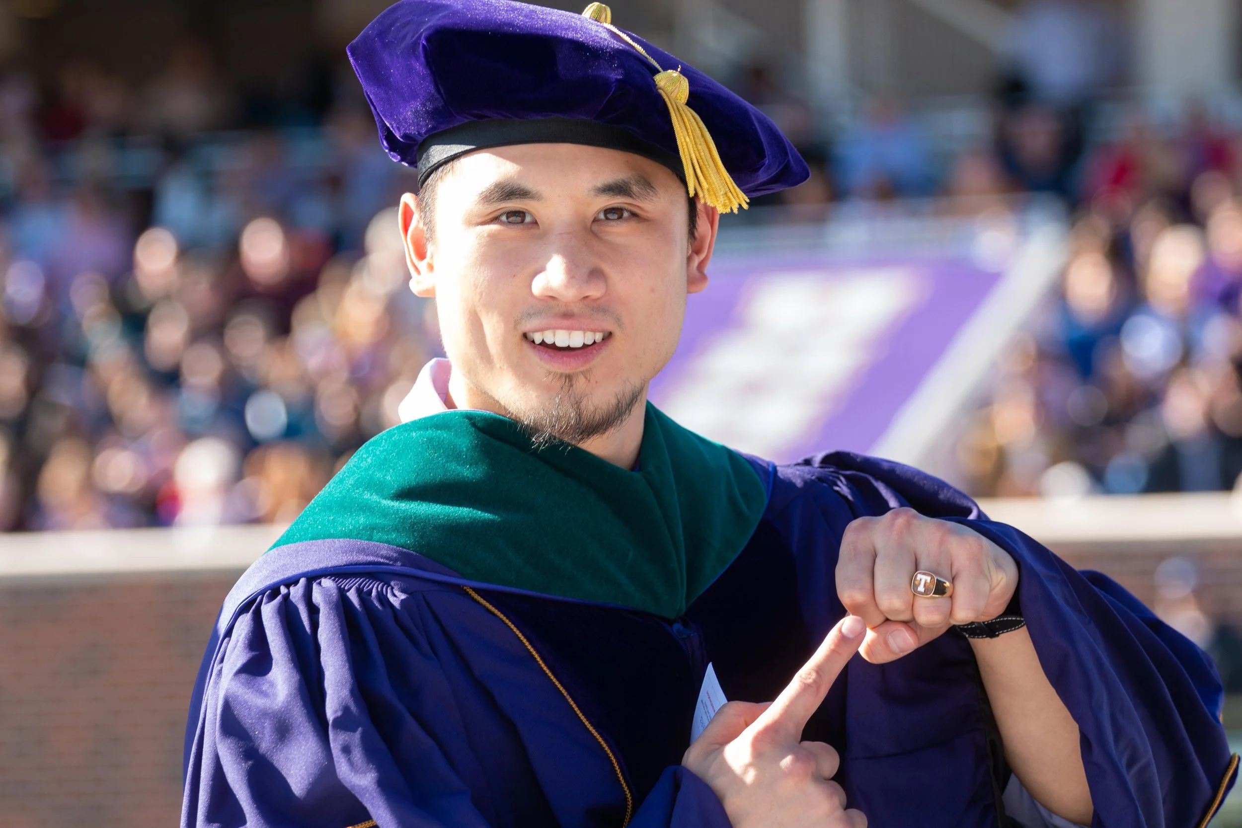 A young man in graduation cap and gown at a graduation ceremony, pointing to his ring.