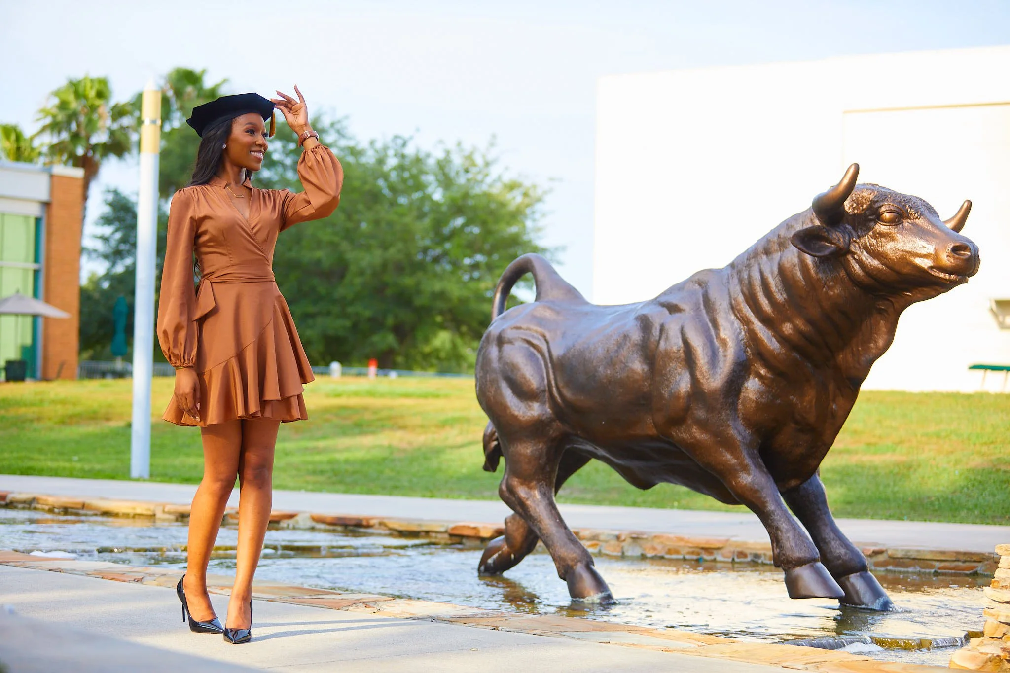 A woman in a brown dress and black heels wearing a graduation cap, smiling and adjusting her cap, standing near a bronze bison statue in an outdoor park setting.