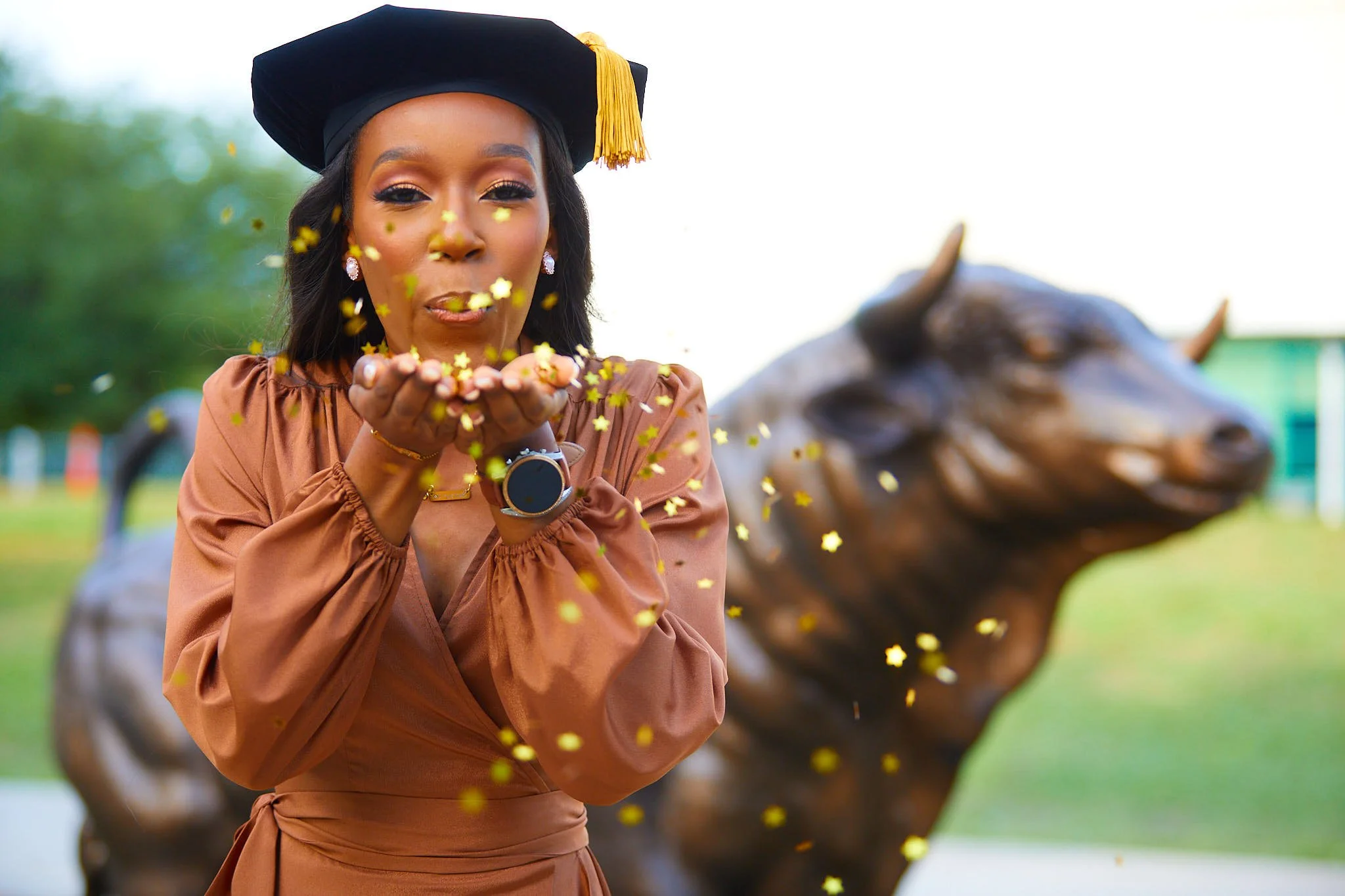 A woman wearing a cap and gown blowing golden star-shaped confetti in front of a bull statue outdoors.