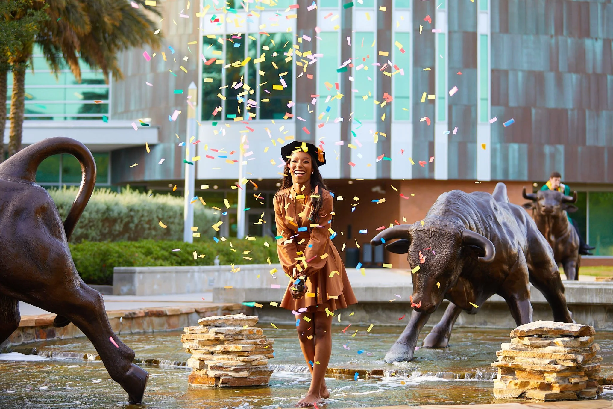 A woman in a brown dress celebrating graduation with confetti at a fountain with bronze animal sculptures, including a buffalo in front and a bison in the background, outside a modern building.
