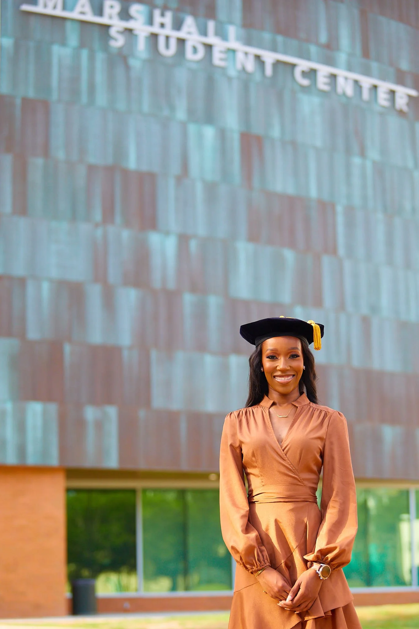 A woman in a tan dress wearing a graduation cap standing in front of a building with a sign that reads 'Marshall Student Center'.