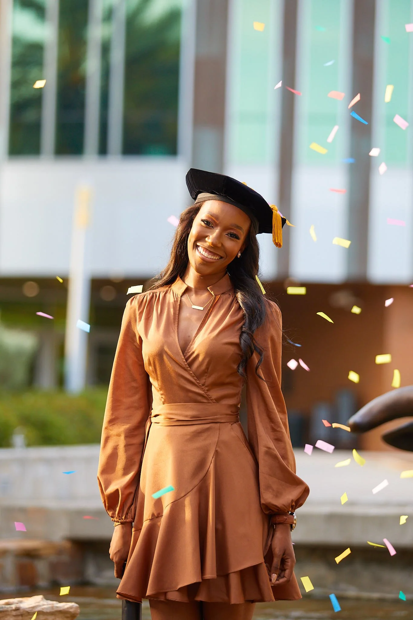 Young woman in brown dress and graduation cap with yellow tassel celebrating graduation with confetti falling around her outside.