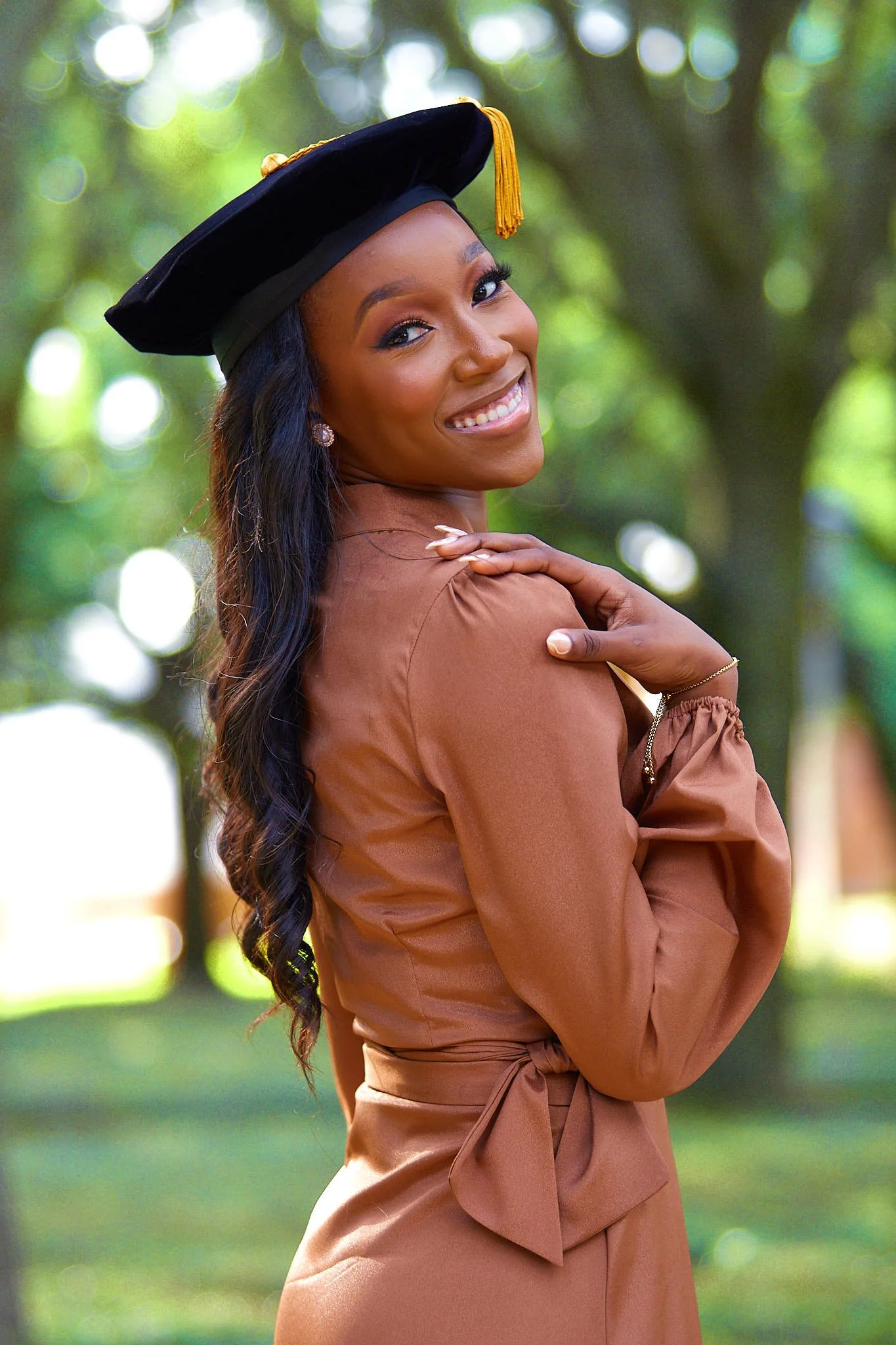 A young woman in a cap and gown, smiling outdoors during graduation, with a blurred green background and bokeh lights.