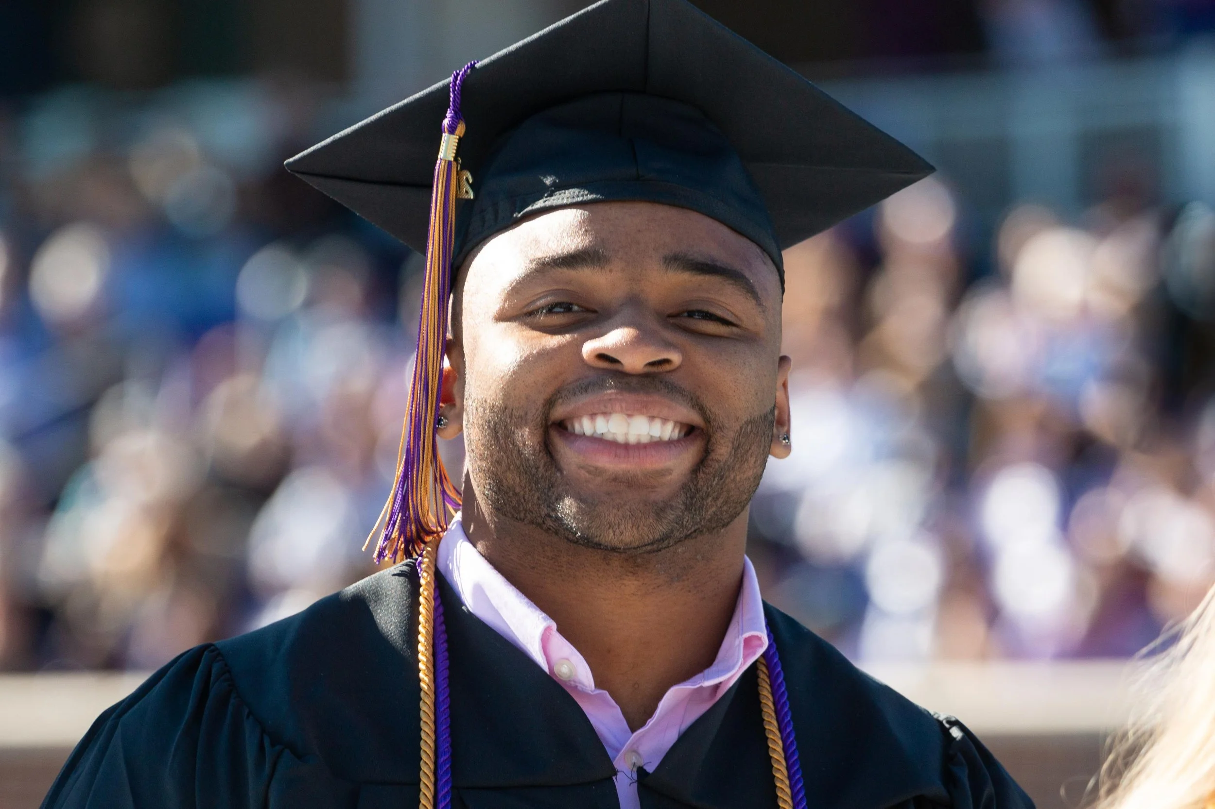 Graduation cap and gown worn by a smiling young man at an outdoor graduation ceremony.