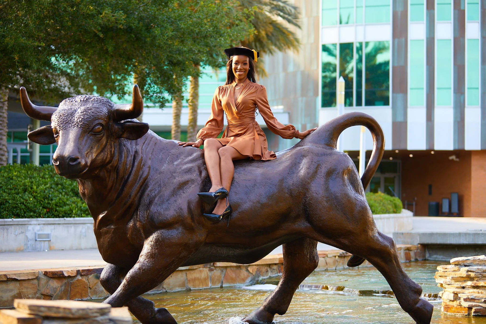 Woman in a brown dress and black heels sitting on a large bronze bull statue in an outdoor urban setting with modern glass buildings and trees in the background.