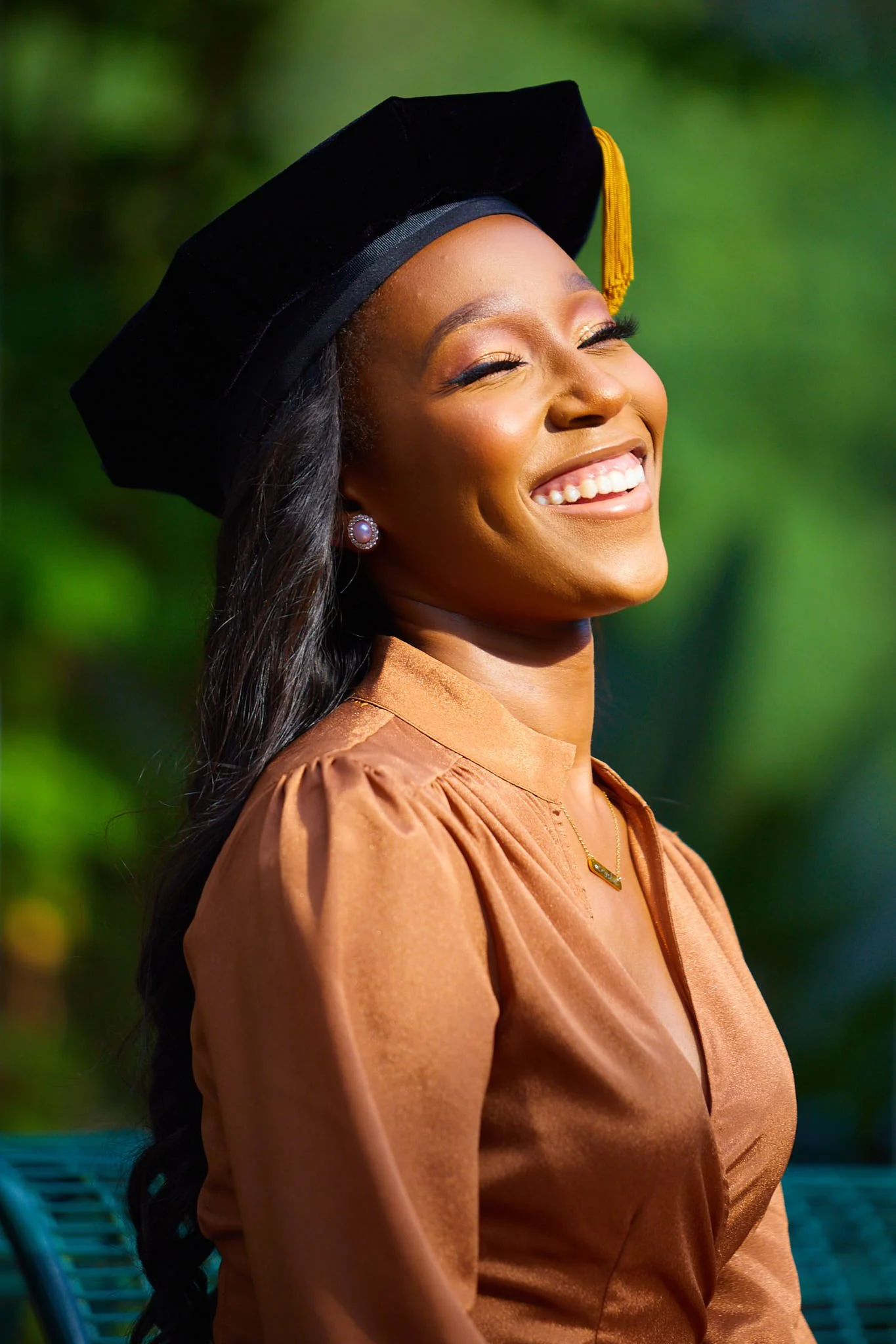 A smiling woman dressed in a graduation cap and gown in an outdoor setting with green foliage in the background.