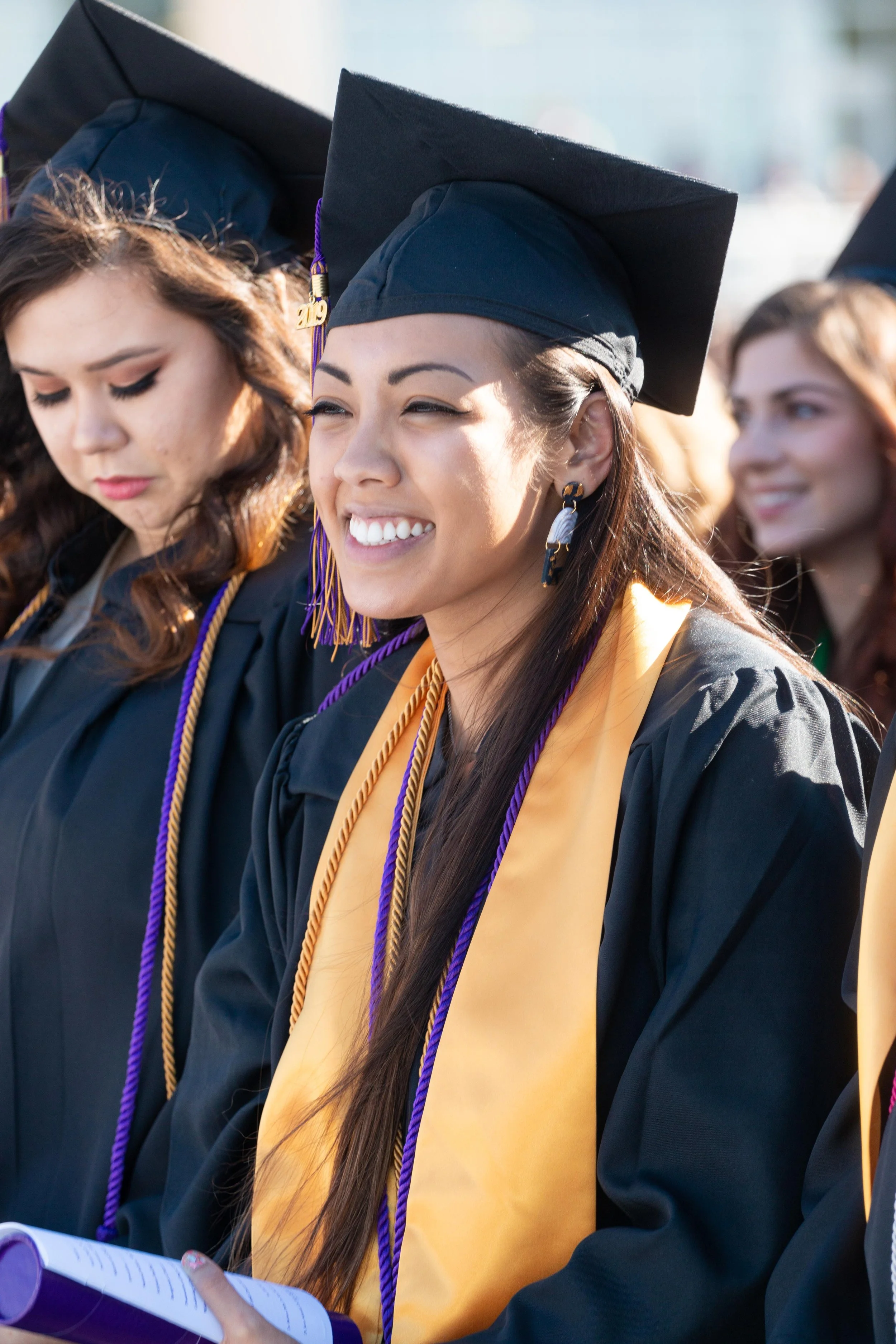 Group of diverse female students in graduation caps and gowns at an outdoor graduation ceremony, some smiling and others looking down.