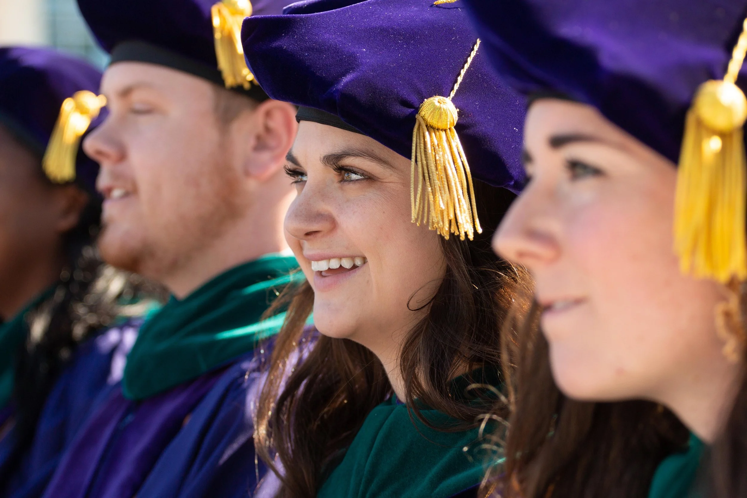 Graduates wearing purple caps with gold tassels and green robes, standing in a line, smiling during graduation ceremony.