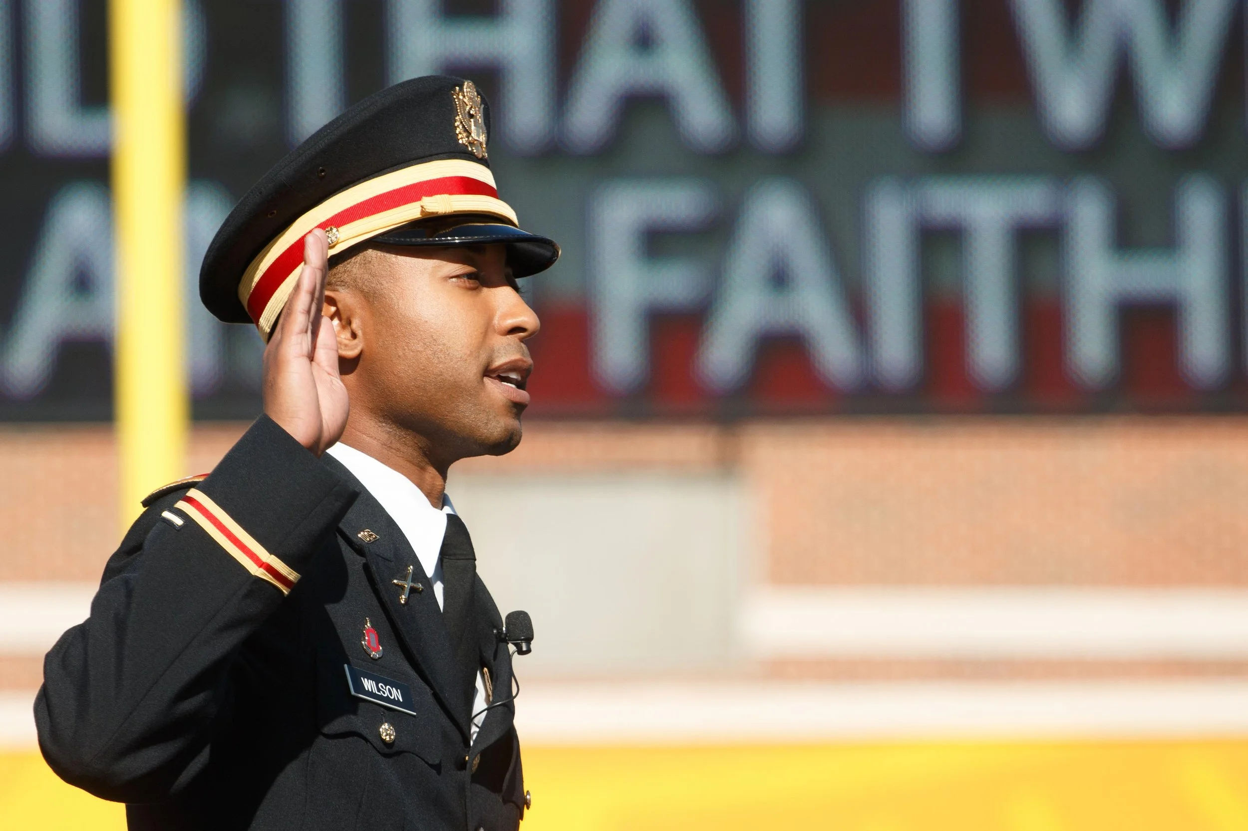 A uniformed officer participating in a formal ceremony, raising his right hand in a salute or pledge, with a blurred background of a stadium or sports field.