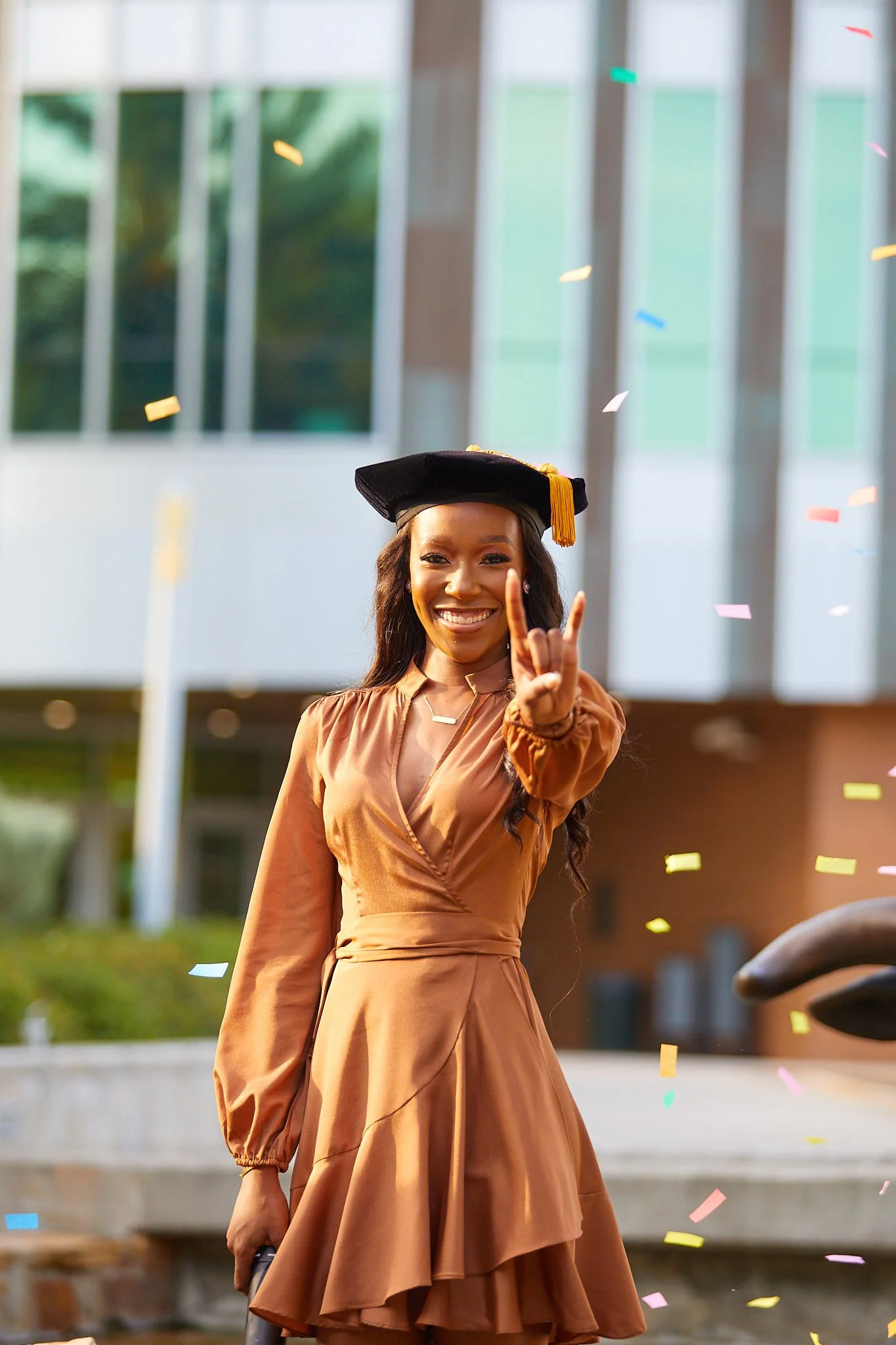 A young woman in a brown dress and graduation cap smiling and holding up her hand in a peace sign, with colorful confetti falling around her outdoors near a modern building.