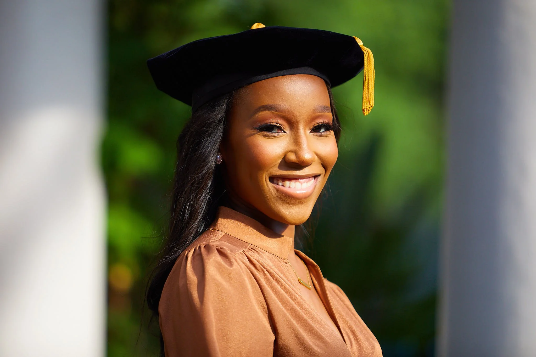 A young woman in graduation attire, wearing a black cap with a gold tassel, smiling outdoors with blurred greenery in the background.