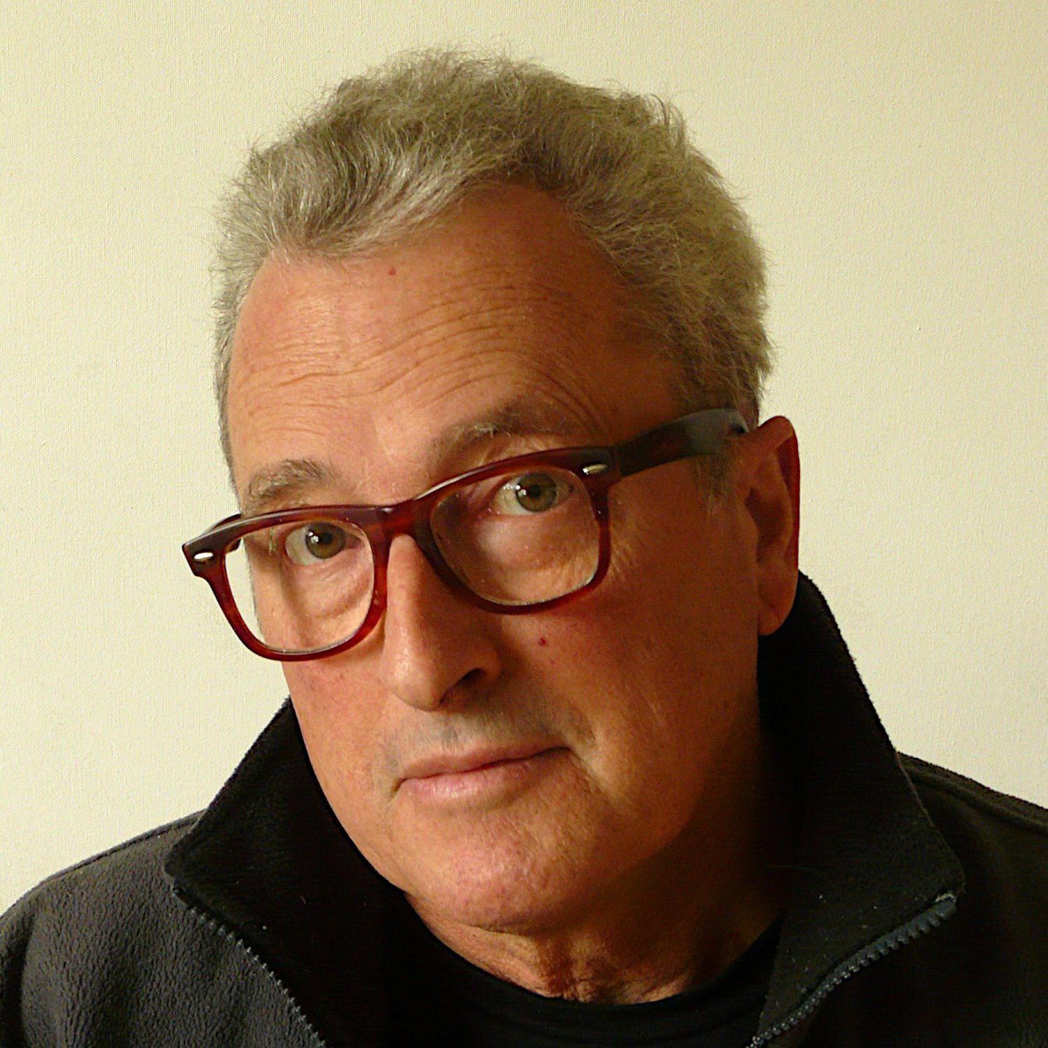 Close-up of contemporary artist Peter Greyson with gray hair, wearing red glasses and a black fleece jacket against a plain light background.