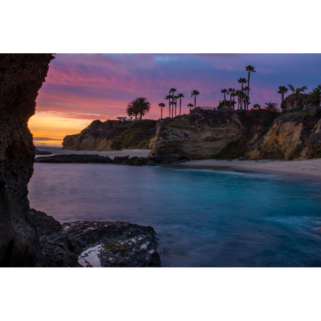 Laguna Twilight – Montage Point dramatic colorful sky with palm trees rocky coastline and crystal clear turquoise waters revealing sand and rocks in Laguna Beach California