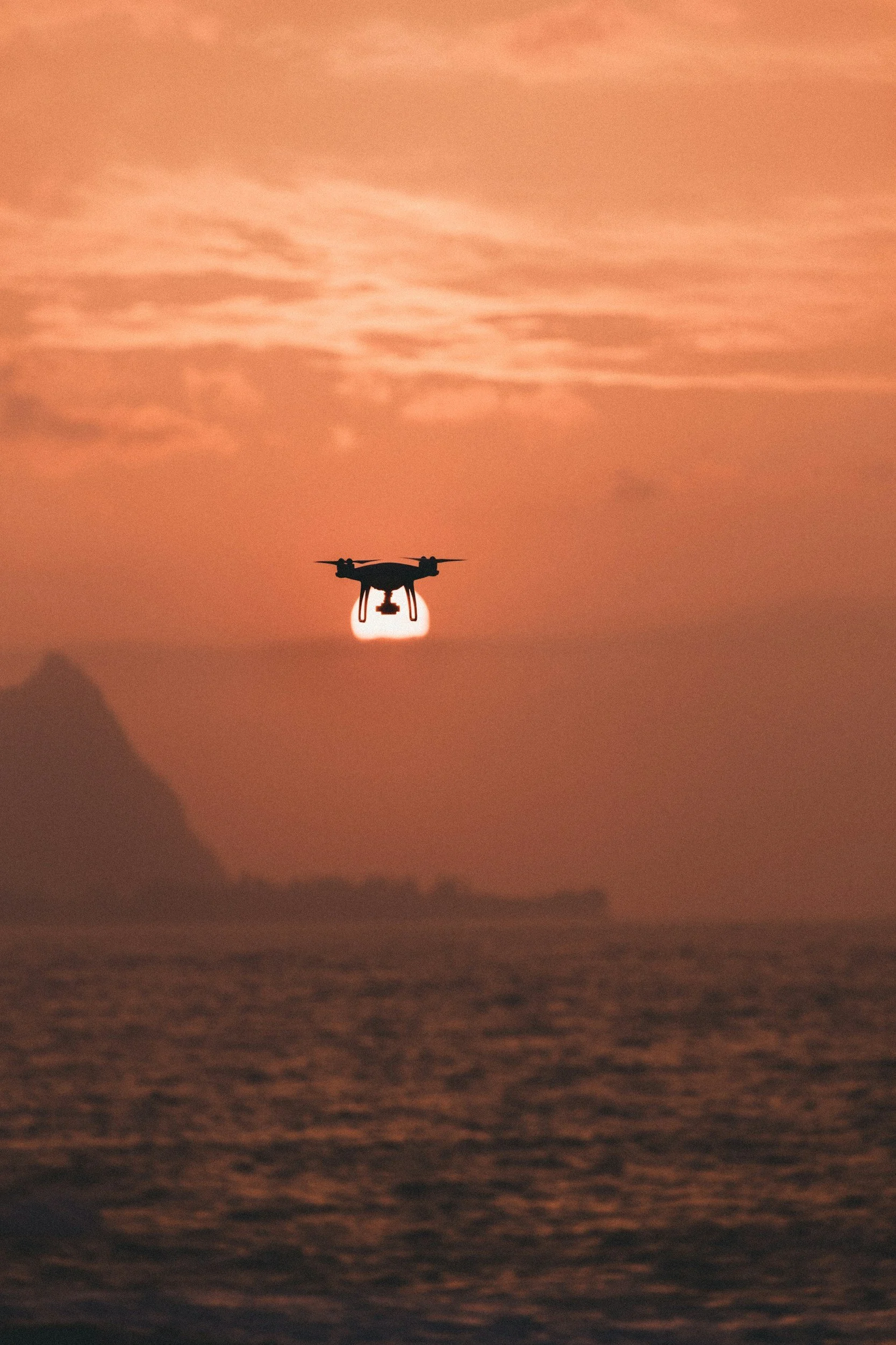 A drone flying over the ocean at sunset, with the sun low on the horizon and clouds in the sky.