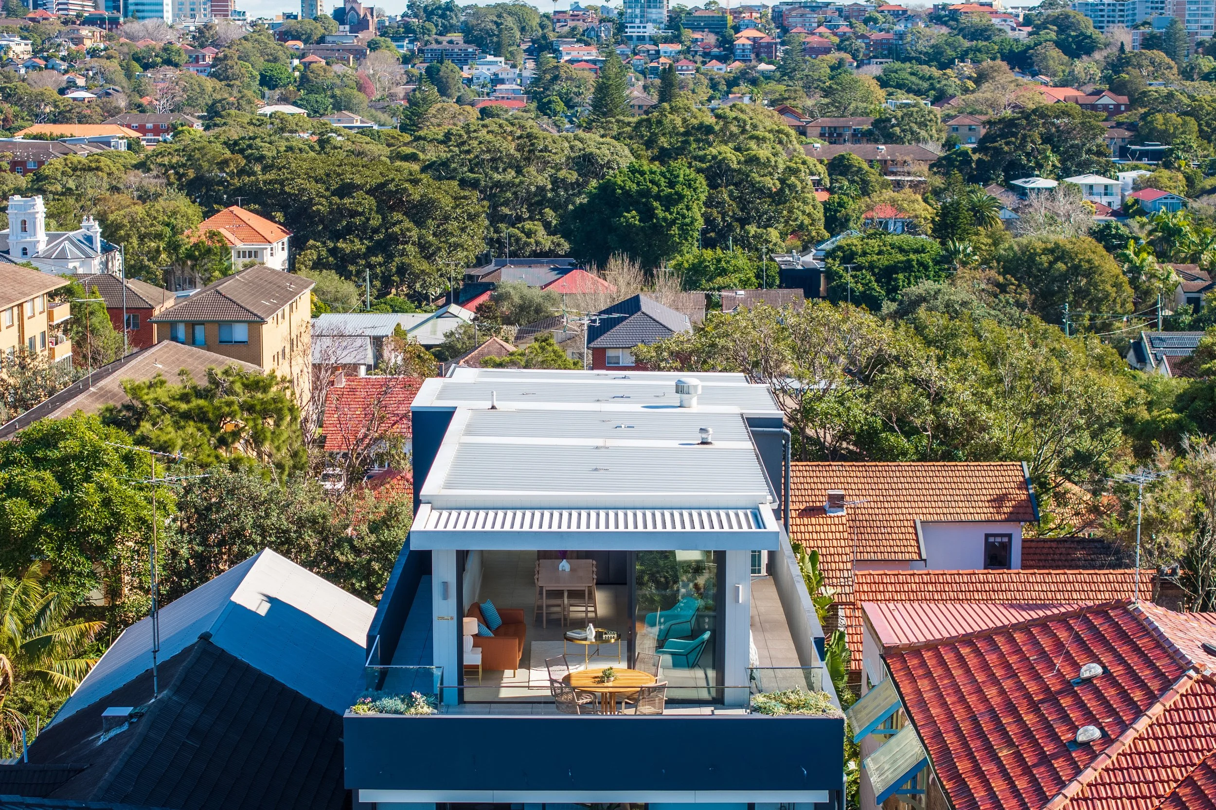 An aerial view of a residential neighborhood with a modern rooftop terrace in the foreground, surrounded by trees and colorful houses under a clear blue sky.