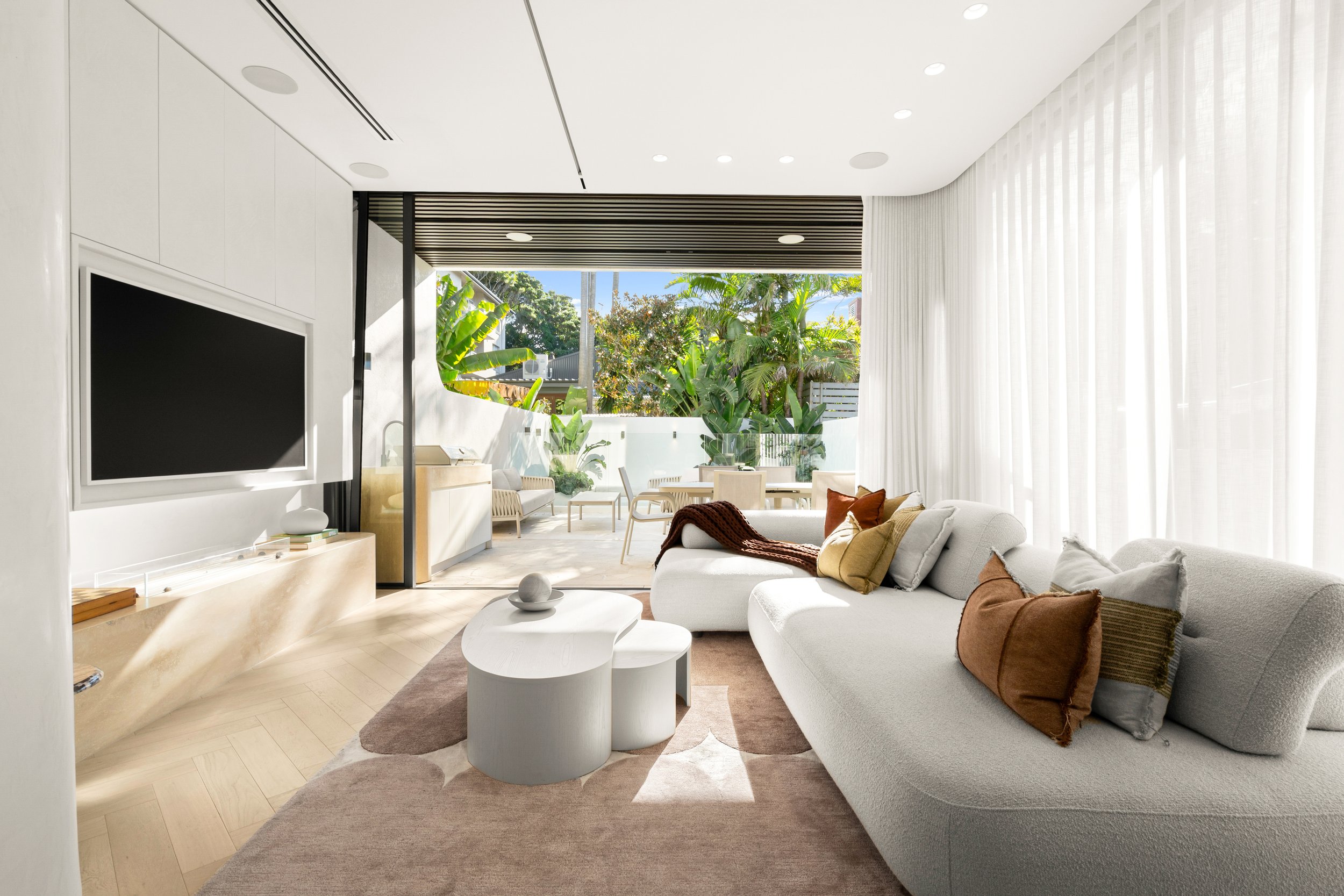 Modern living room with white sofa, brown cushions, white coffee table, beige rug, large glass doors with sheer curtains, outdoor patio with green plants, and a view of blue sky and trees