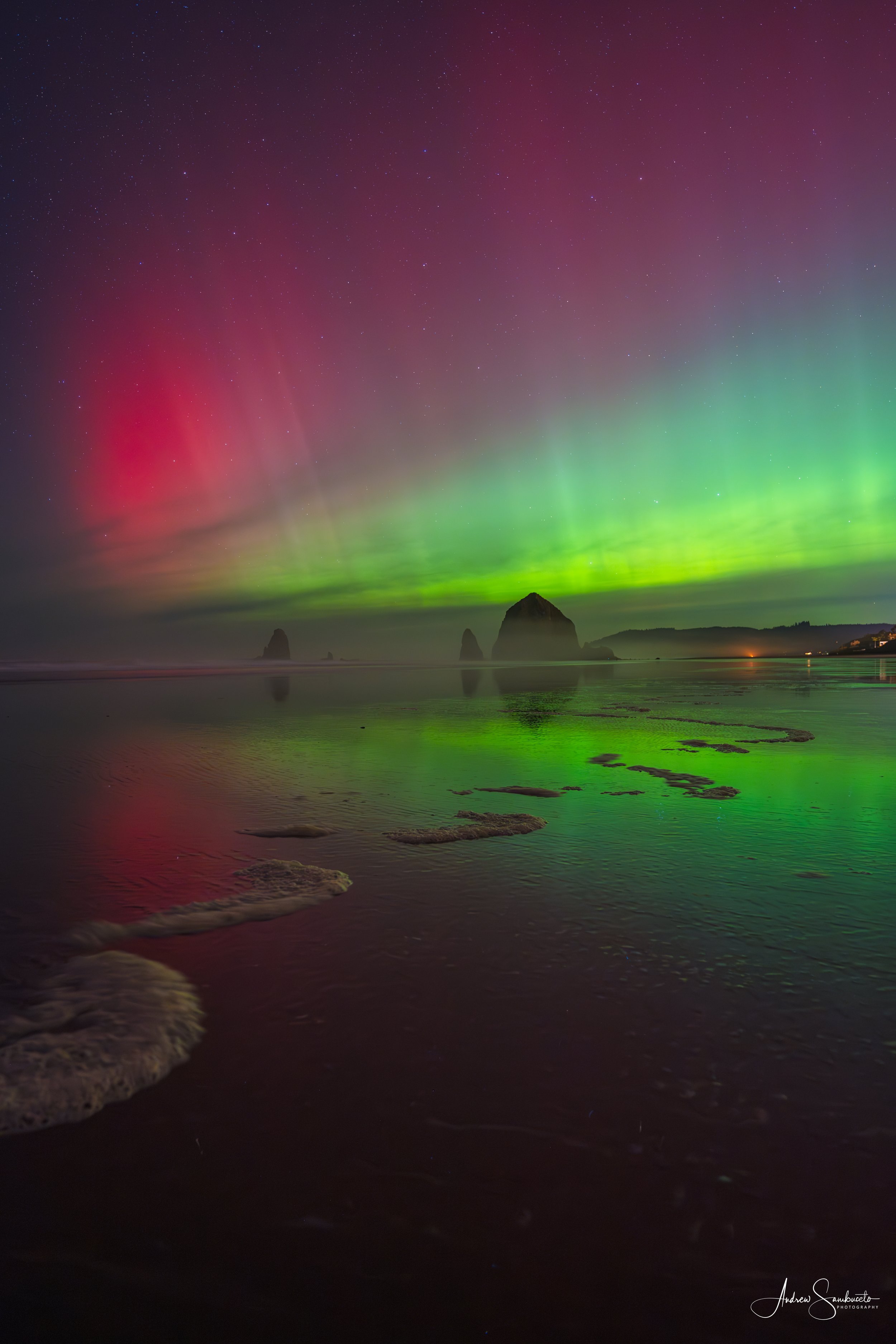Northern lights over a beach with rocks and ocean, starry night sky in the background.