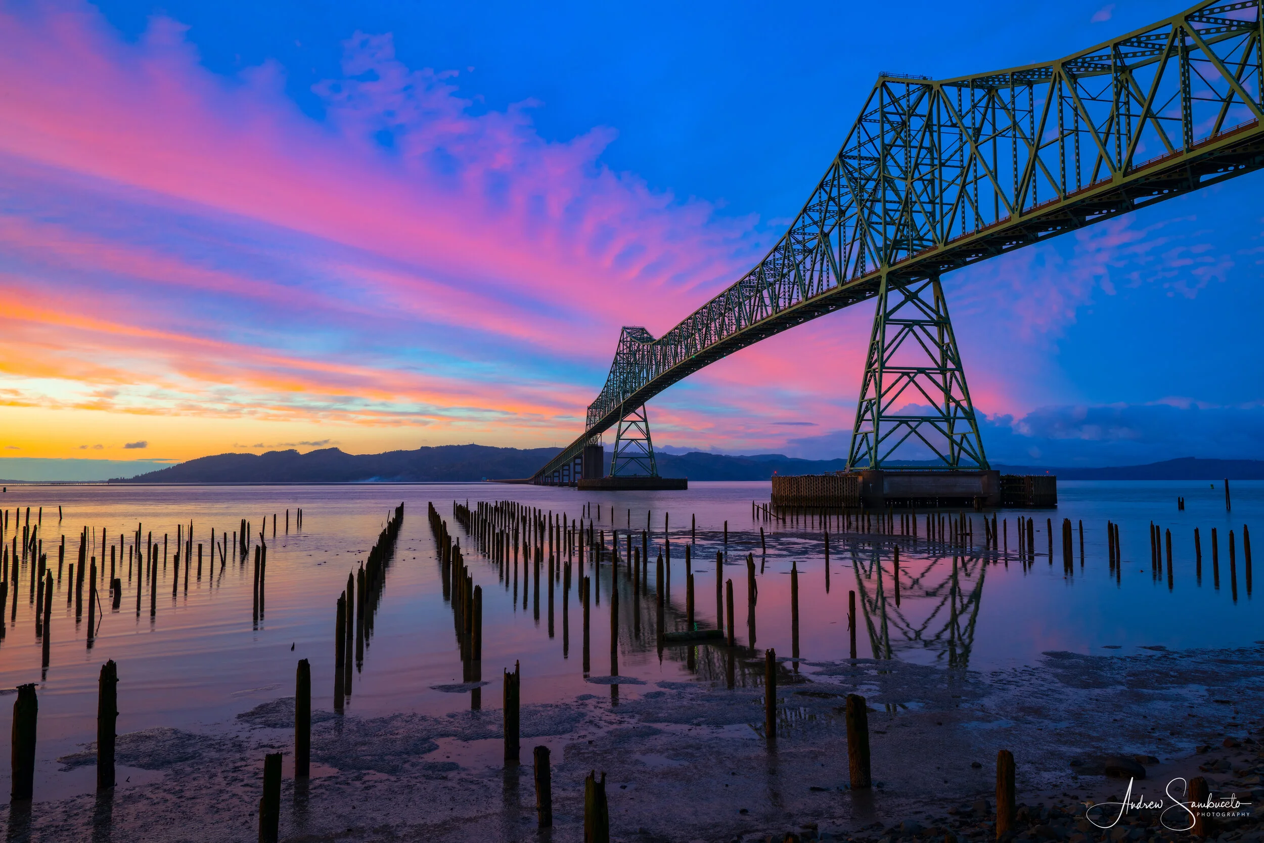 Colorful sunset over a large steel bridge spanning across a body of water, with wooden posts in the foreground and mountains in the distance.