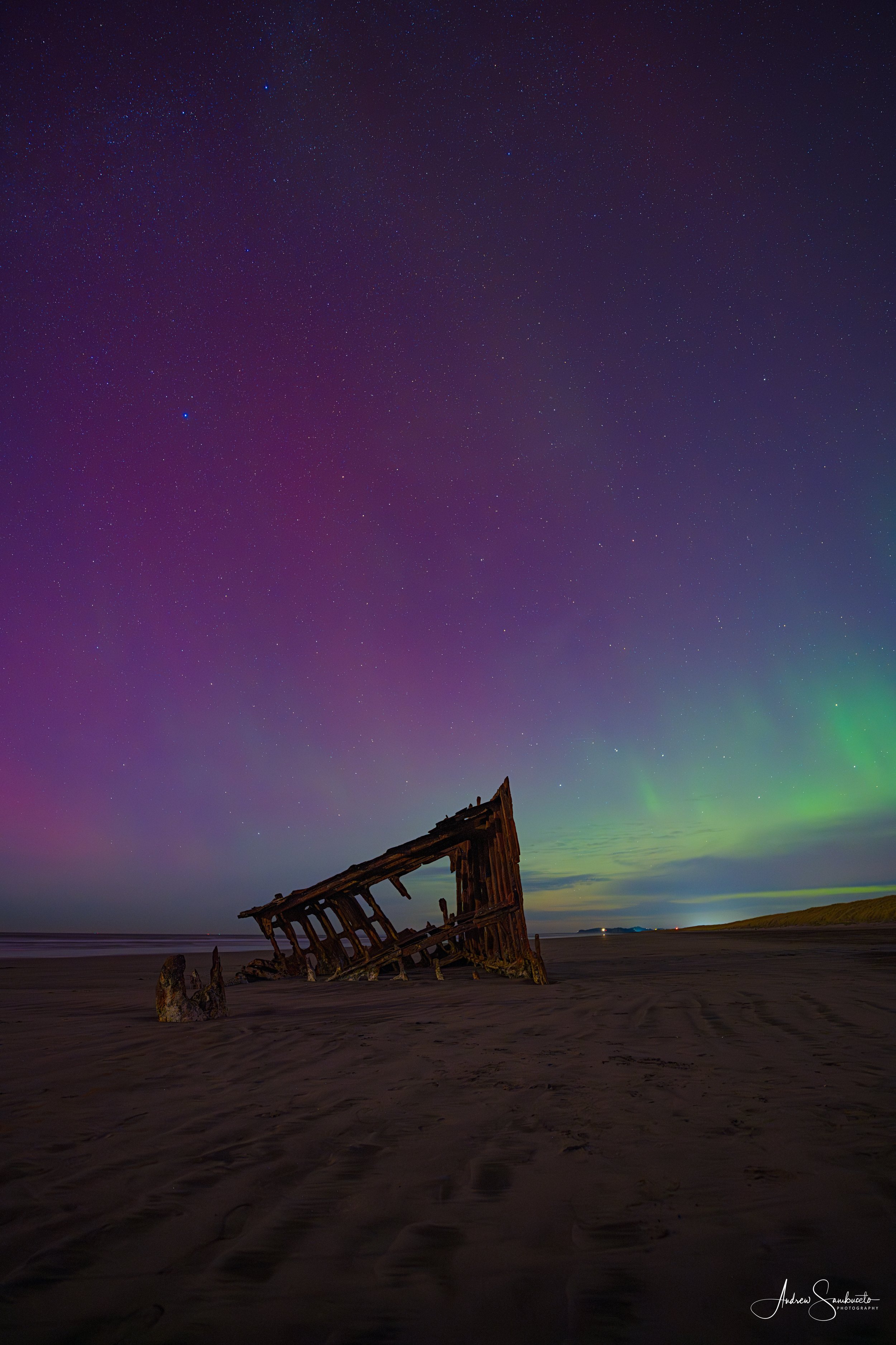 Nighttime shot of a sandy beach with an old, dilapidated shipwreck partially buried in the sand. The sky is filled with stars and the northern lights are visible in the distance, creating a colorful glow.