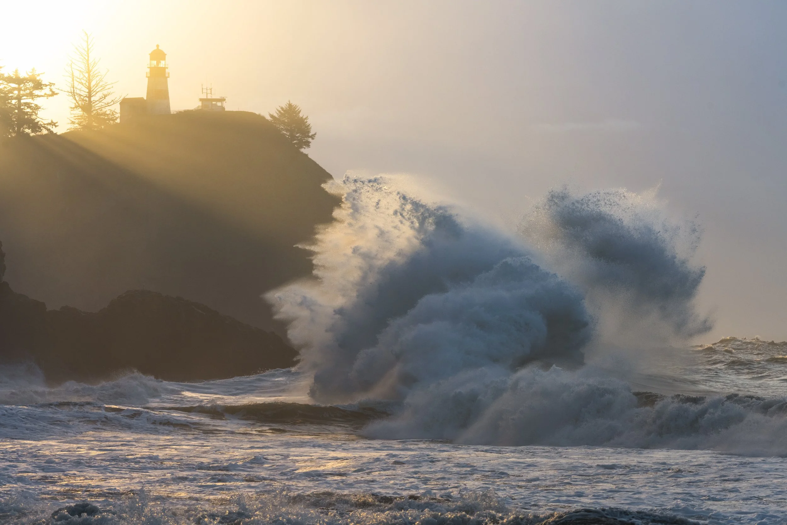 Sunset over ocean waves crashing against rocks with a lighthouse on a cliff in the background.