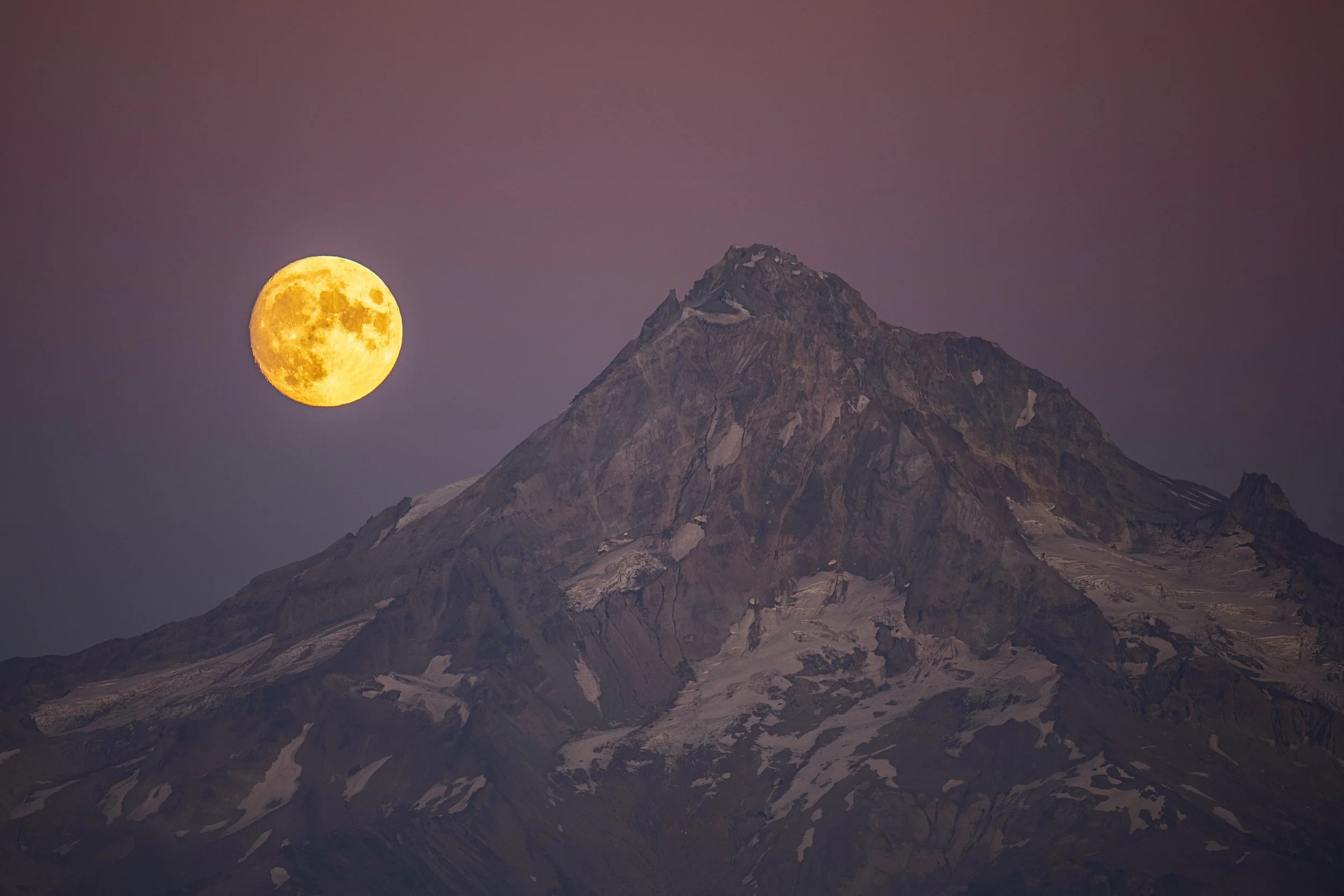 Full moon rising beside a tall mountain with patches of snow and rock, during twilight.