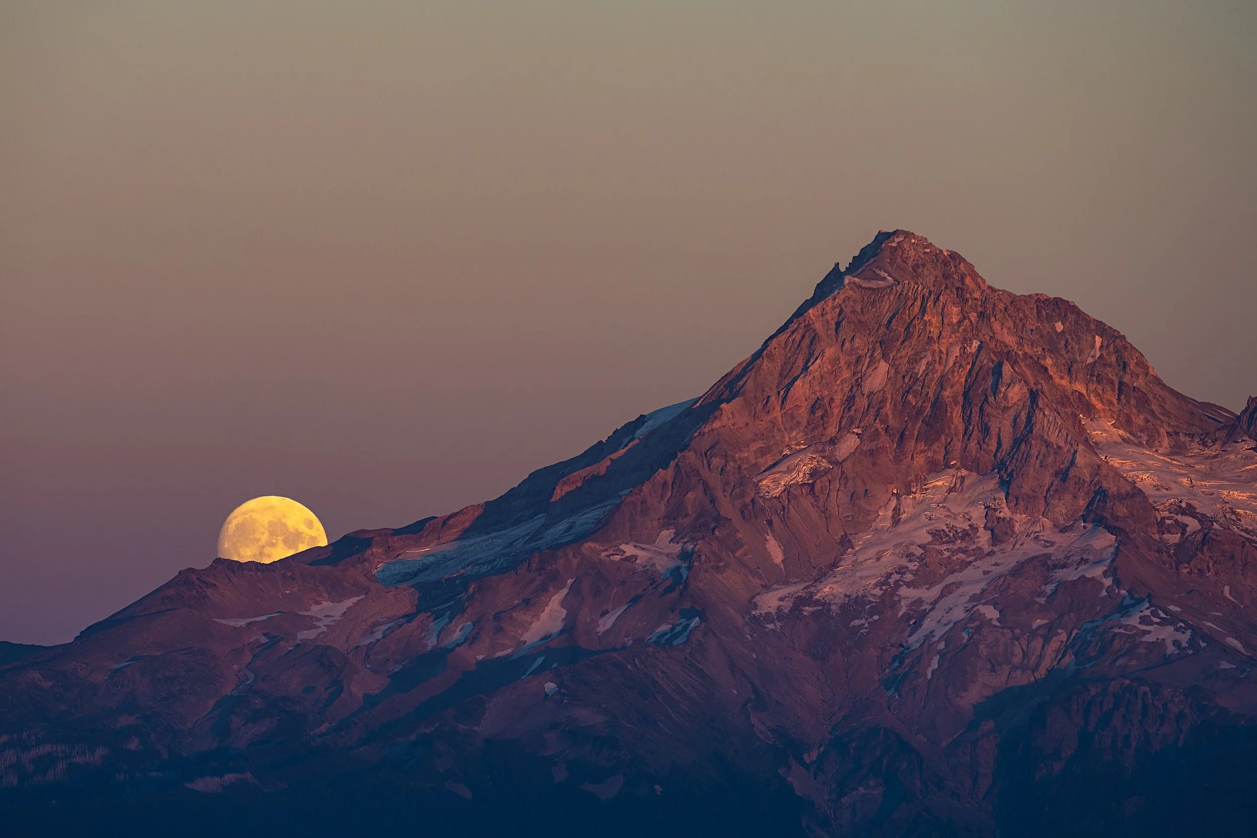 A mountain peak with snow patches at sunset, with a large full moon rising in the sky to the left of the Mt. Hood.