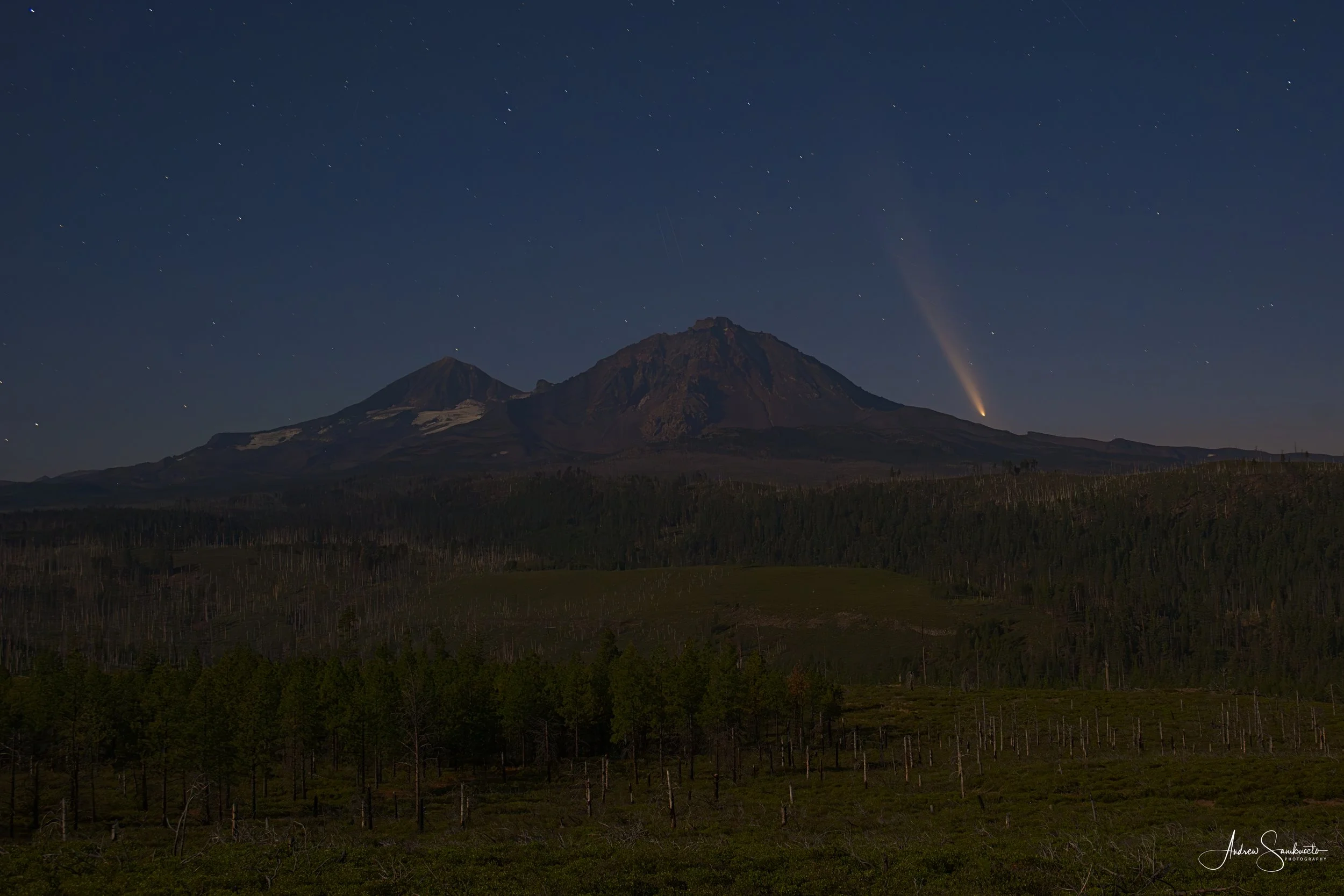 Nighttime landscape of a mountain with a forest in the foreground and a starry sky above, with a bright meteor or comet streaking from the horizon.
