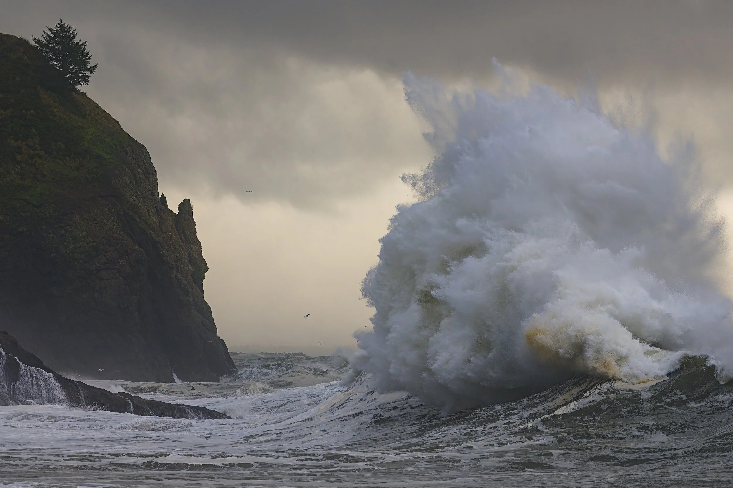 Large ocean wave crashing against rocky cliff with cloudy sky in the background.