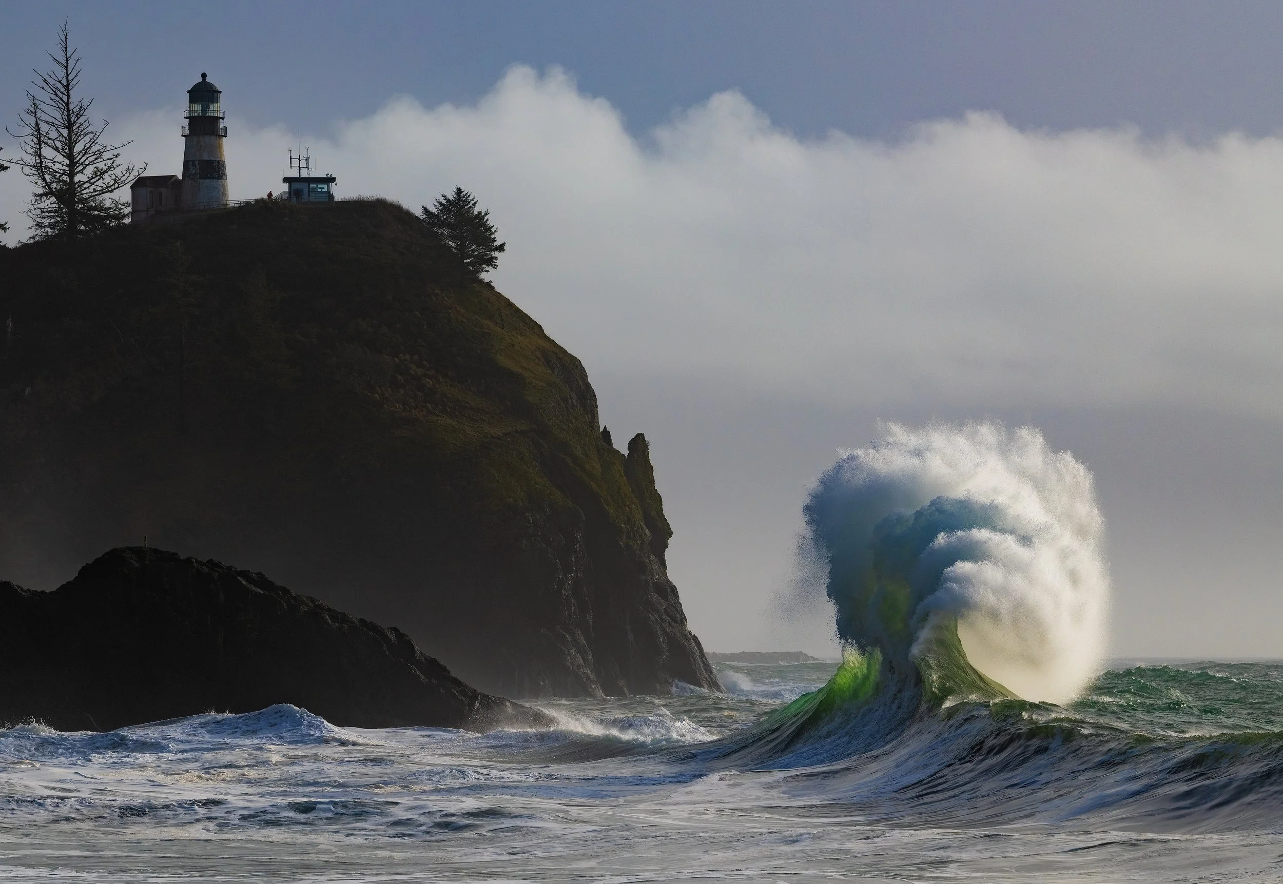 A lighthouse on a cliff overlooks a stormy ocean with large crashing waves.