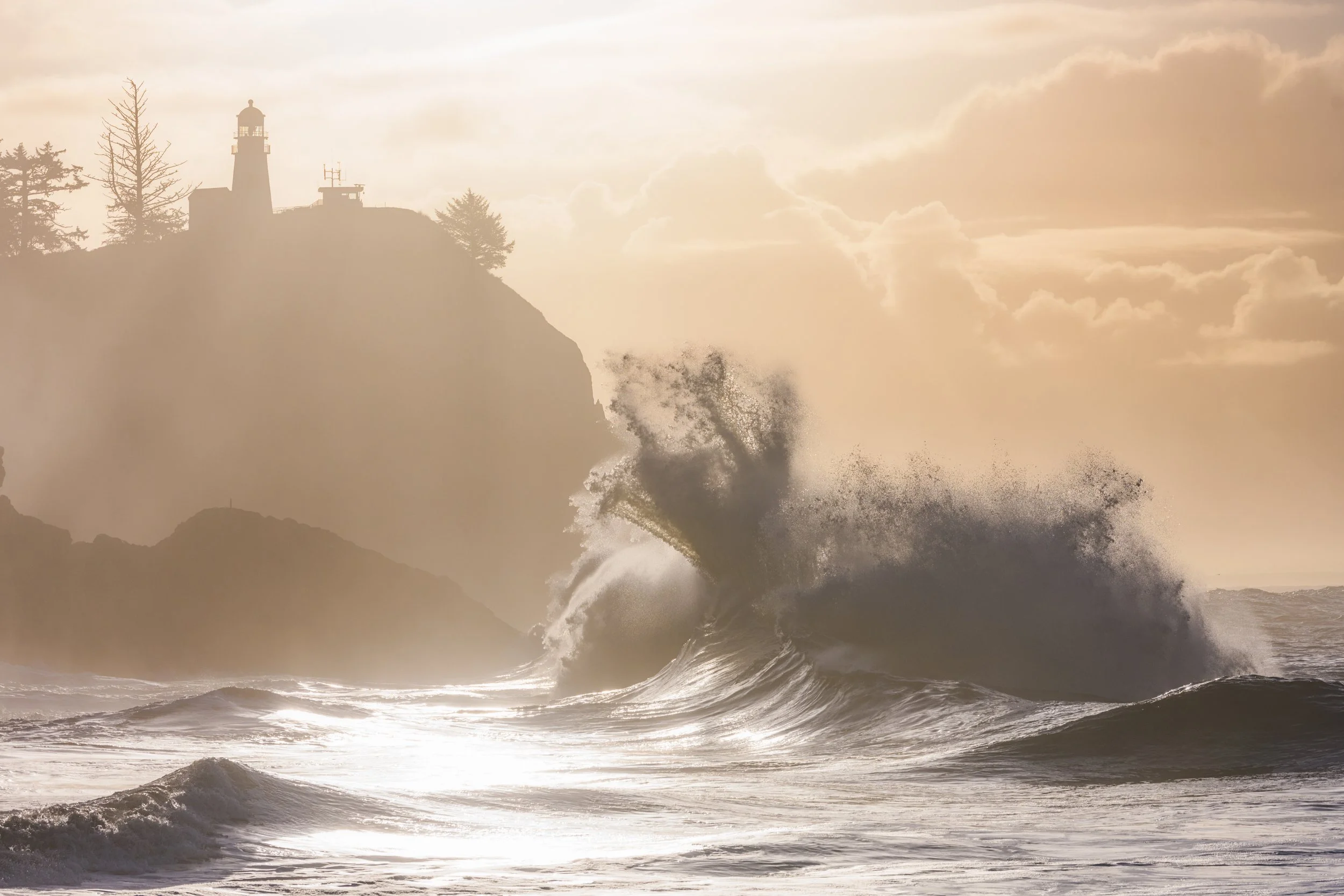 Waves crashing against rocky coastline with lighthouse on cliff at sunset