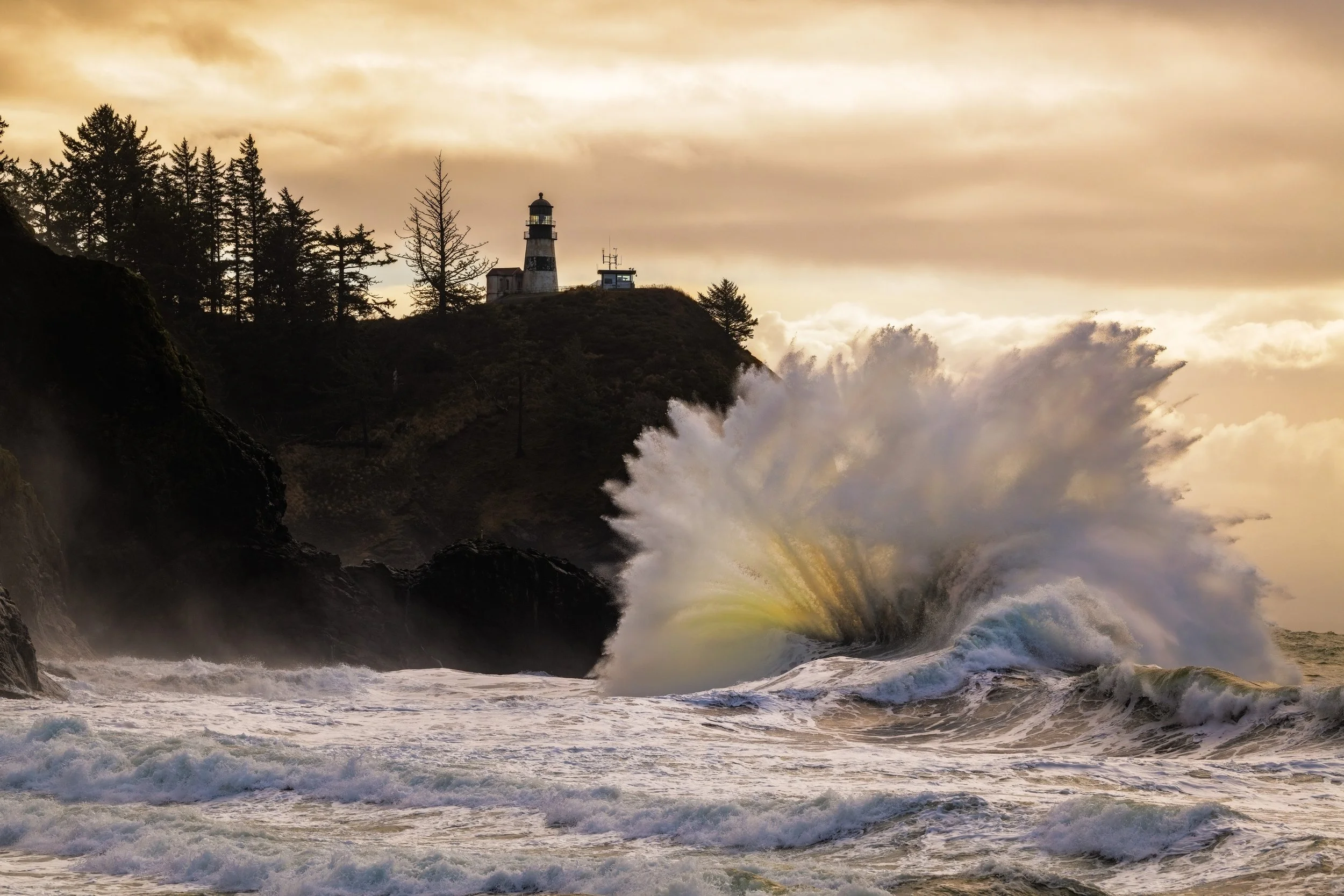 Ocean waves crashing near rocky shoreline with lighthouse on a hill, trees, and cloudy sky at sunset.