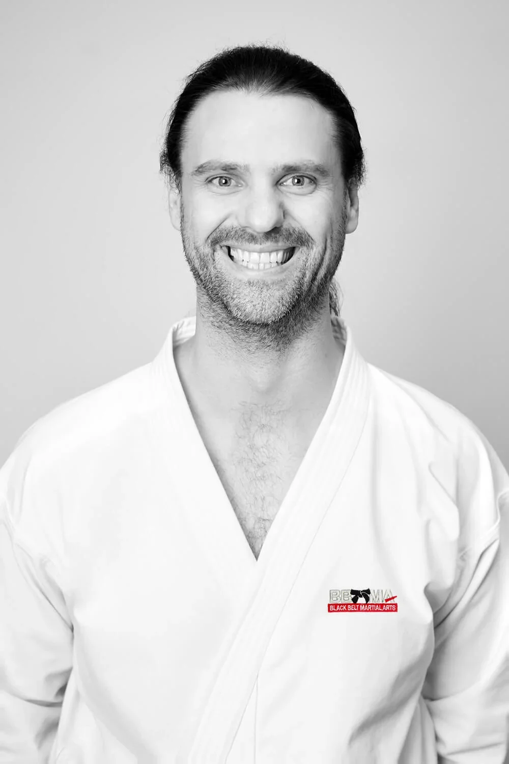 Black and white portrait of a smiling man in a martial arts gi, with long hair and a beard, standing against a plain background.