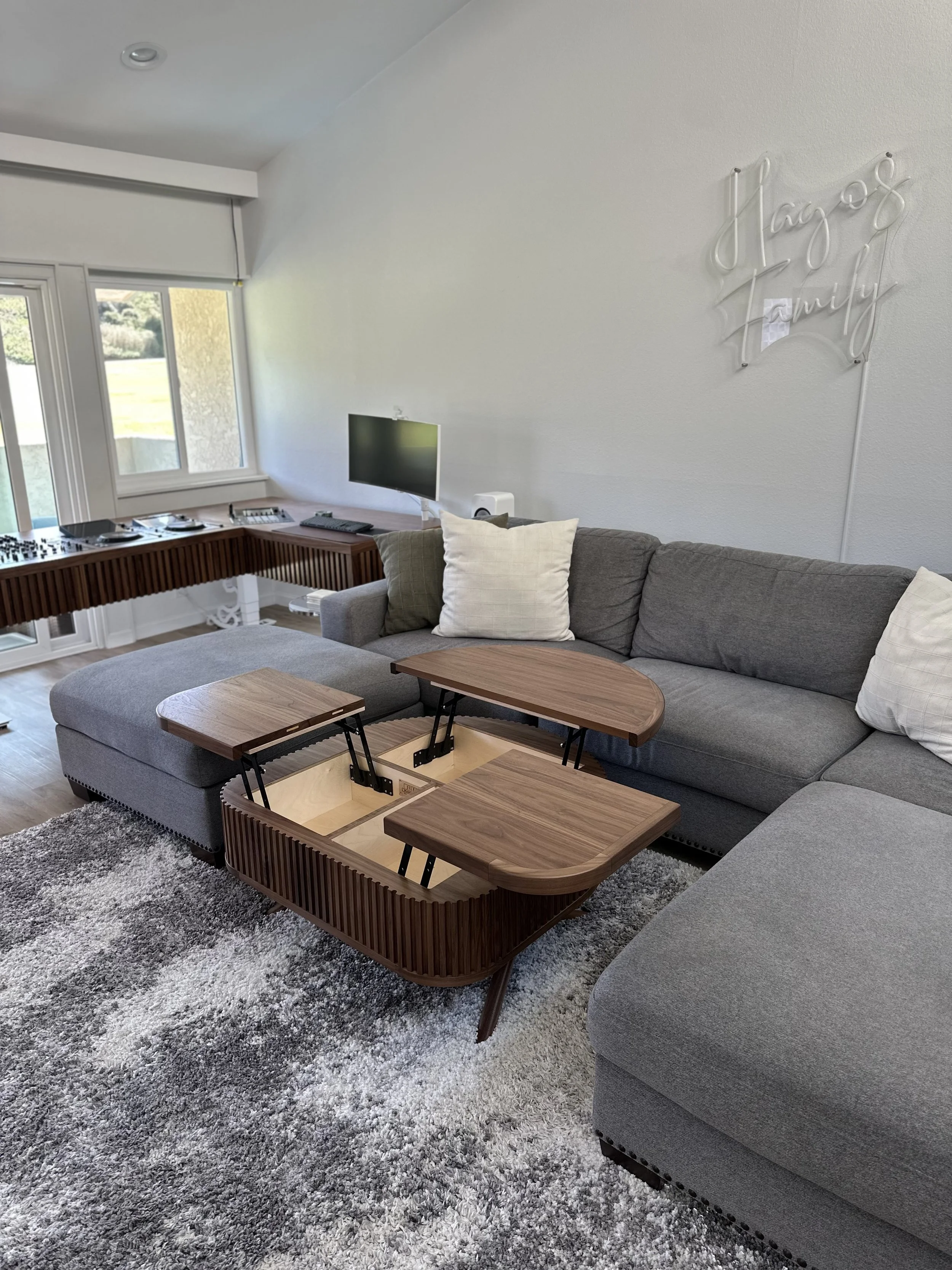 Living room with gray sectional sofa, two wooden nesting tables, a plush gray area rug, a wall-mounted TV, and a wall sign that says "Hugs & Family."