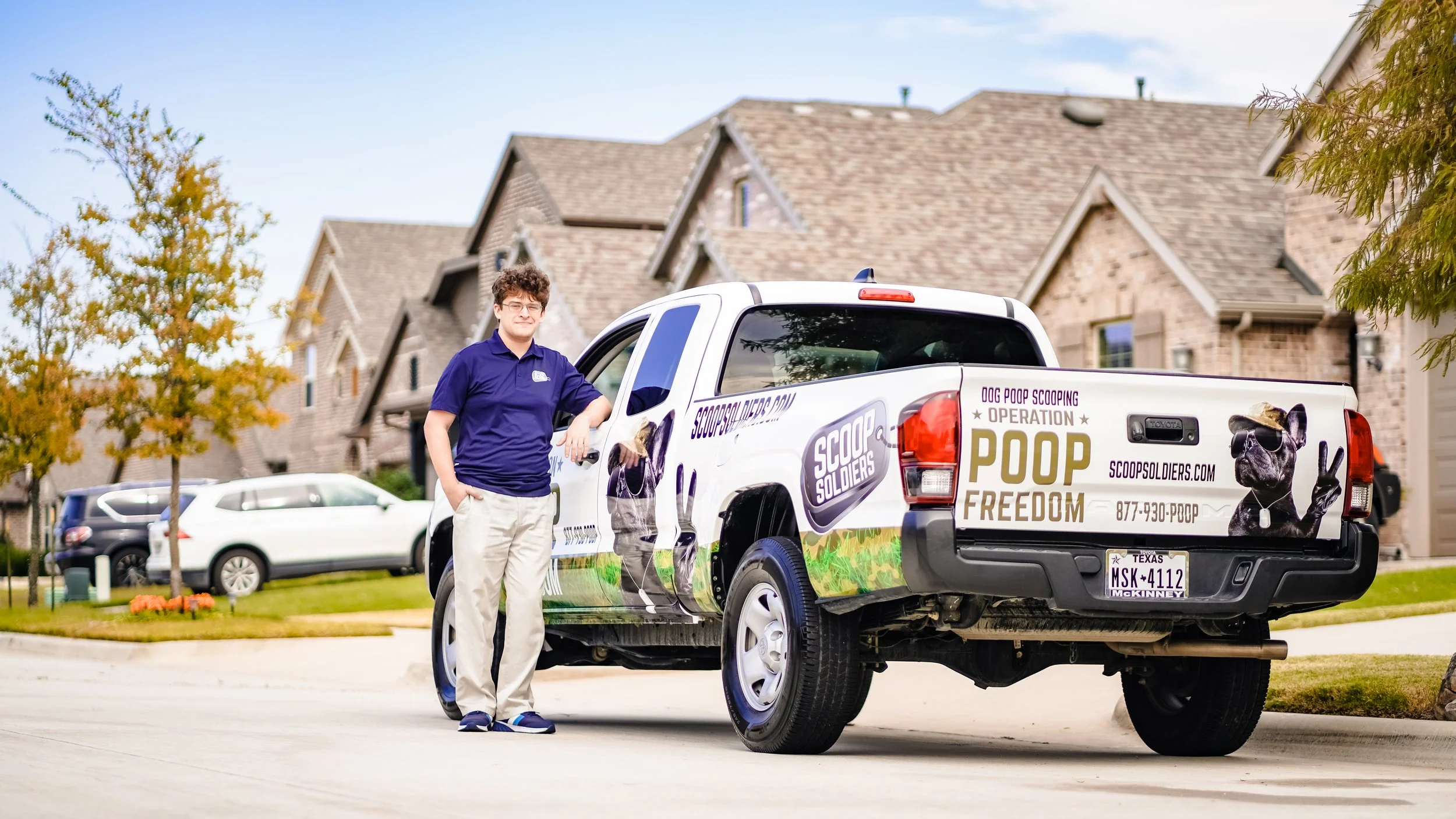 Man standing beside a truck with branding for Scoop Soldiers, a dog poop scooping service, in a residential neighborhood with houses and parked cars.